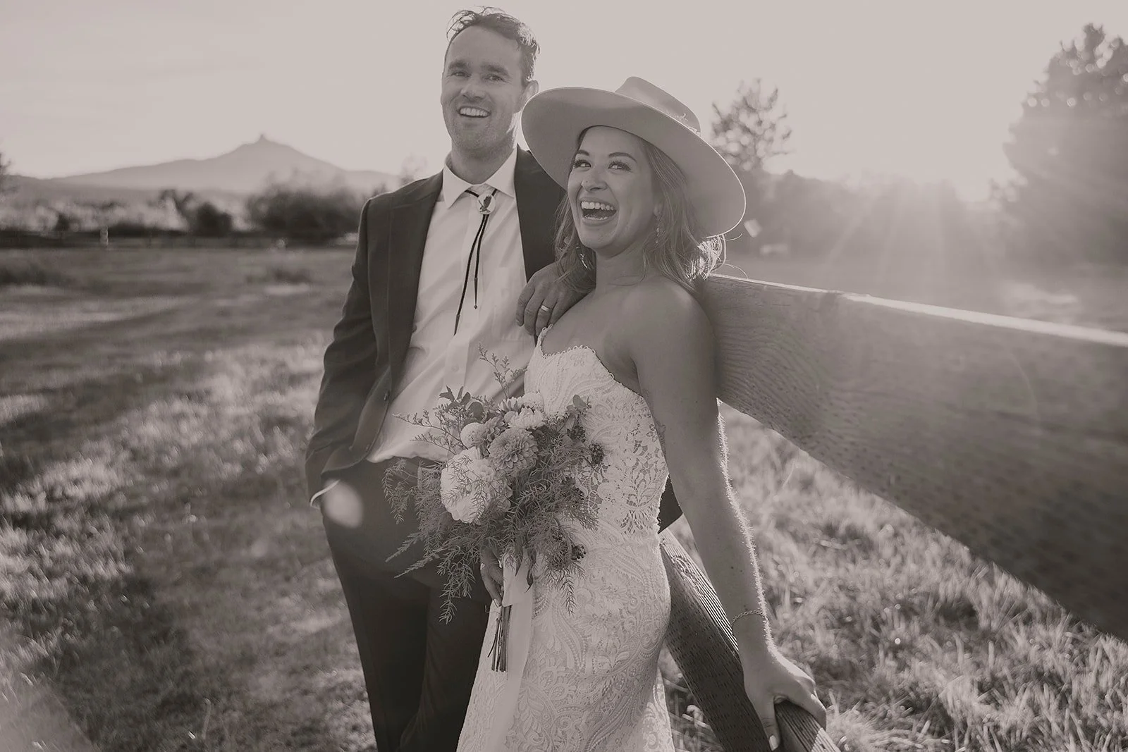 A smiling couple, a man in a suit and a woman in a wedding dress with a large hat, leaning against a wooden fence in a rural outdoor setting during sunset, holding a bouquet of flowers.