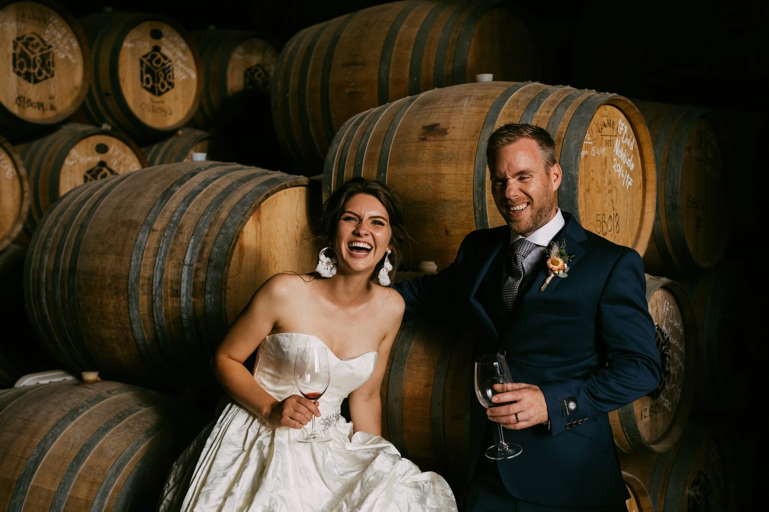 A happy bride and groom in wedding attire smiling and holding wine glasses in a cellar with large wooden barrels in the background.
