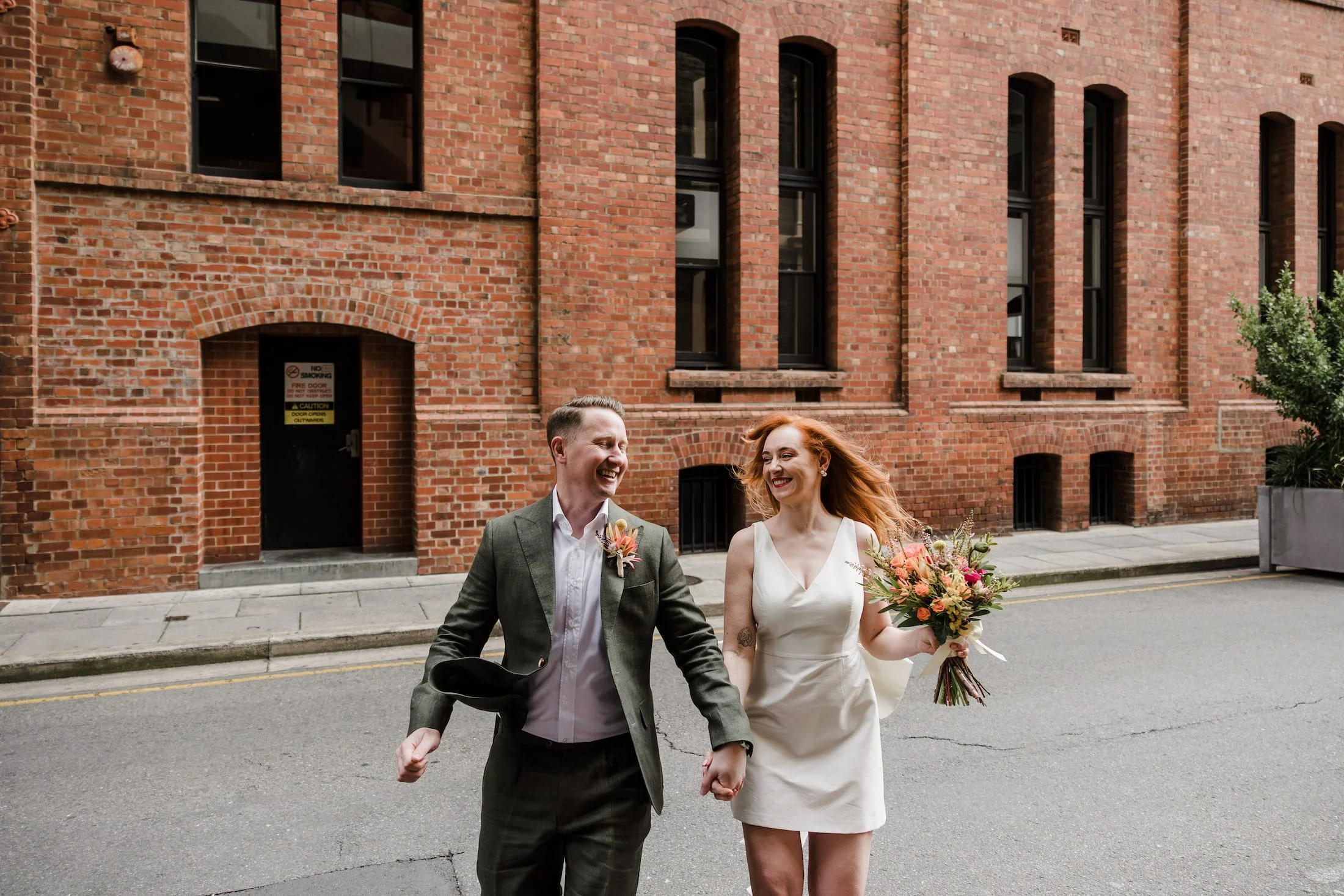 A smiling bride and groom walking hand in hand on a city street in front of a brick building, with the bride holding a bouquet of flowers.