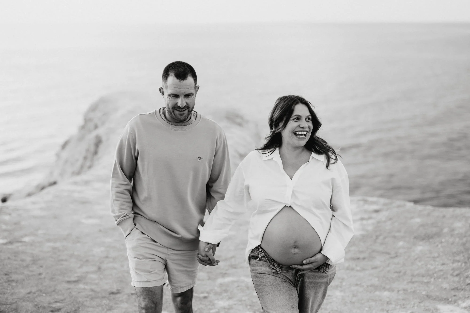 An Adelaide portrait photographer's maternity photo of a couple walking on a beach, holding hands, with the woman visibly pregnant, smiling and enjoying the moment.