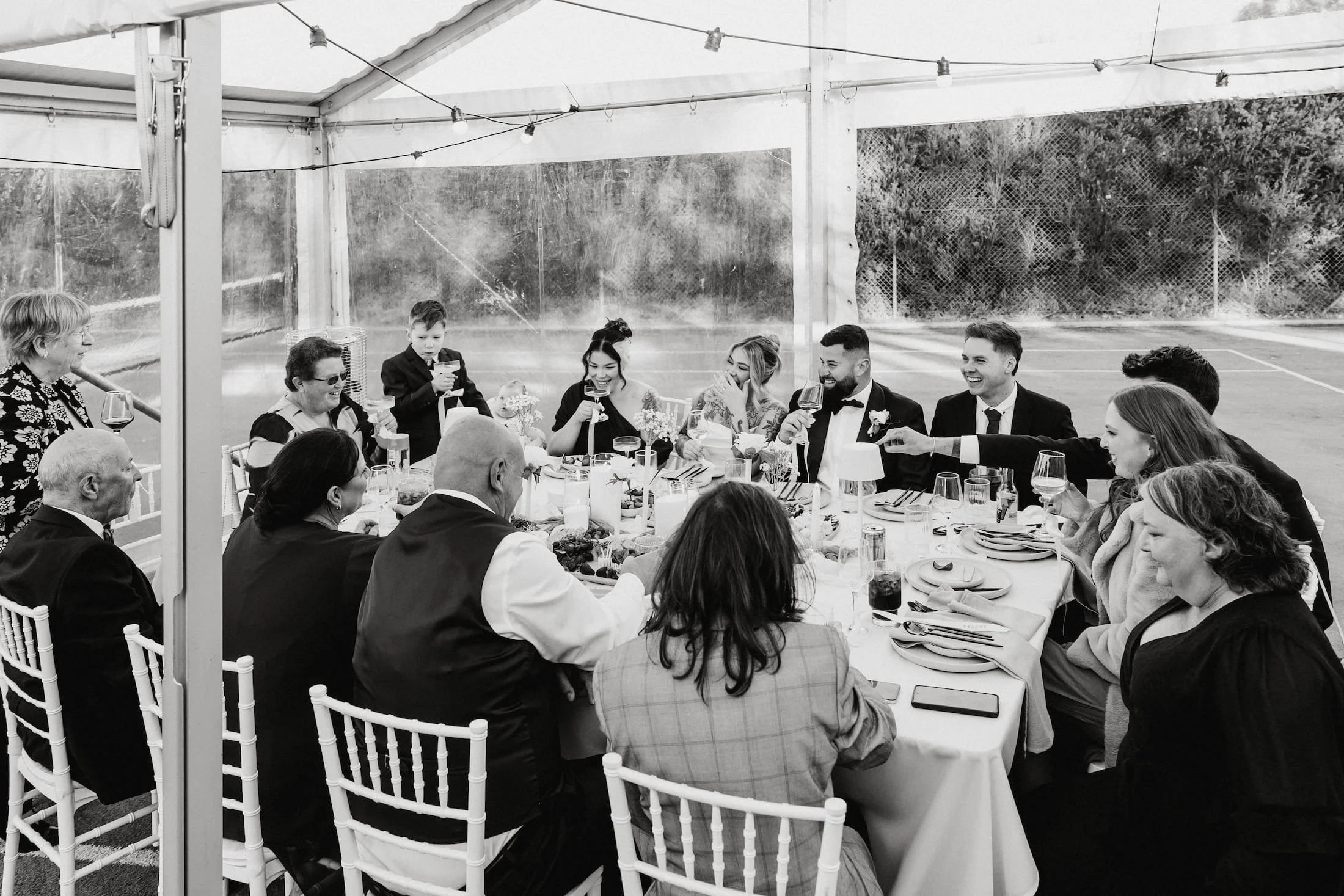 A group of people at a wedding reception seated around a table under a large tent, smiling and raising glasses for a toast.