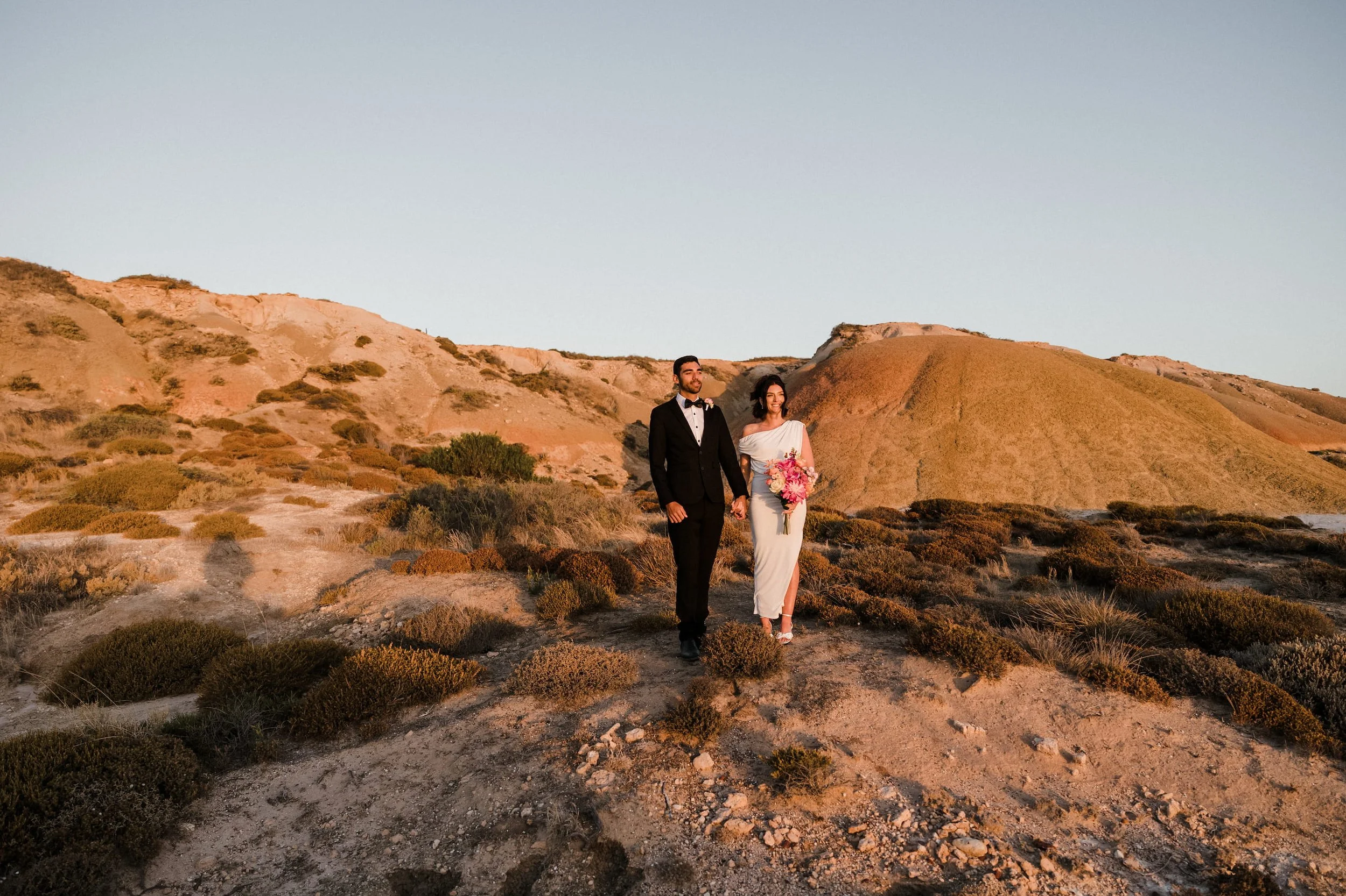 A bride and groom holding hands walking in a desert landscape during sunset, with hills in the background.