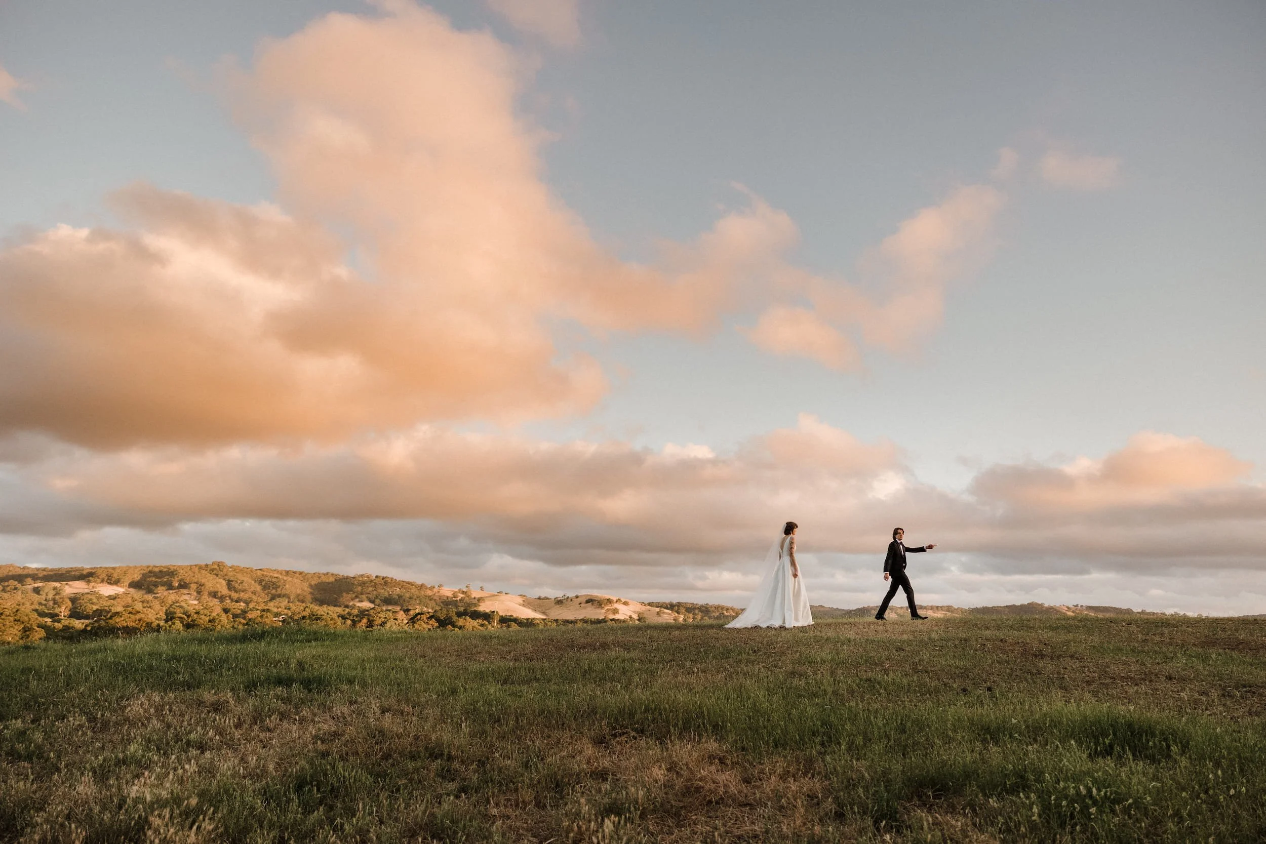 Adelaide wedding photographer Little Car Photography. A couple in wedding attire walking across an open field under a sky with pink and orange clouds at sunset.