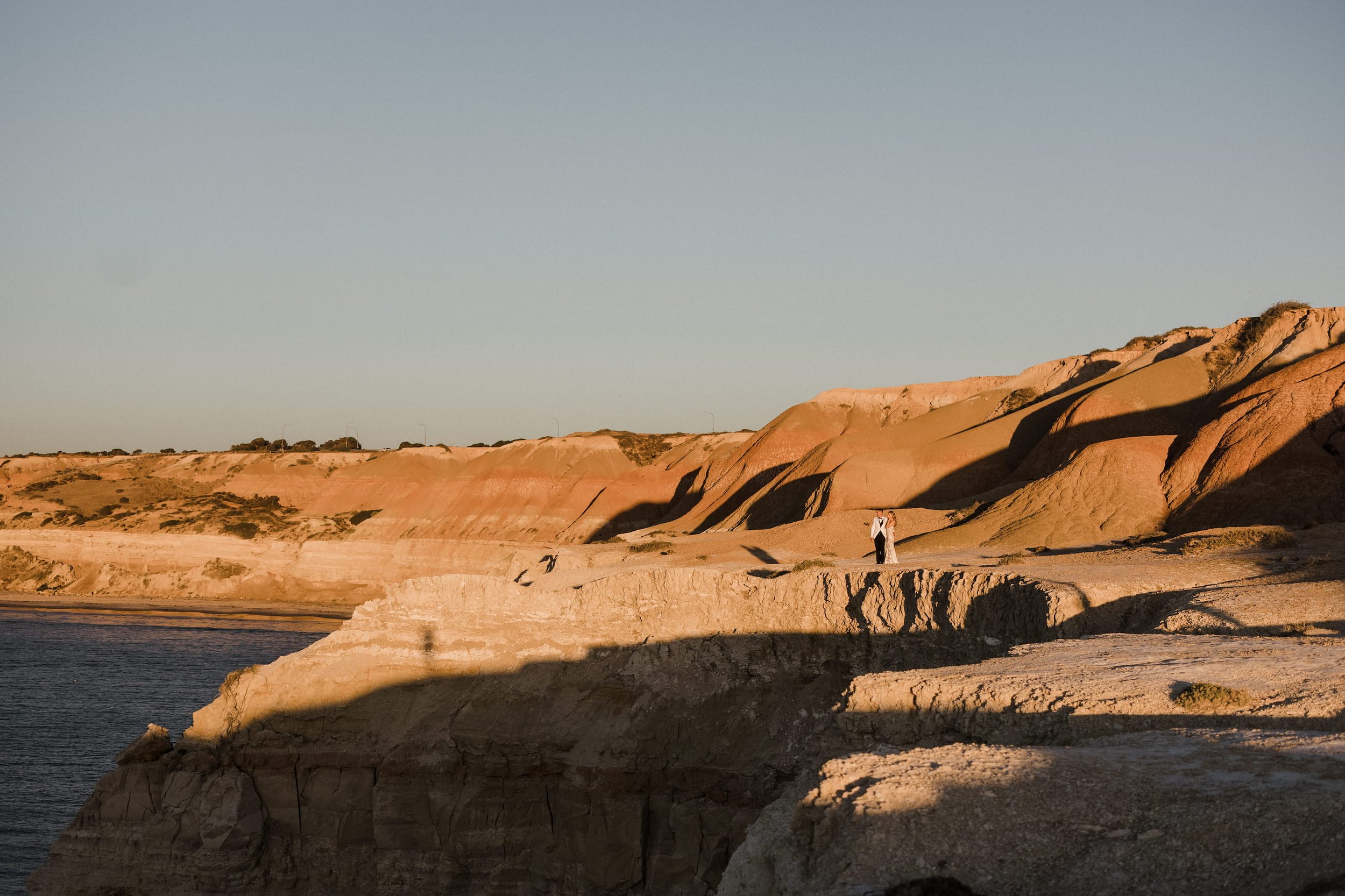 An Adelaide elopement photographer's coastal landscape photograph at sunset featuring layered pink and beige cliffs, a calm body of water in the foreground, and a couple dressed in white standing on the cliffs.