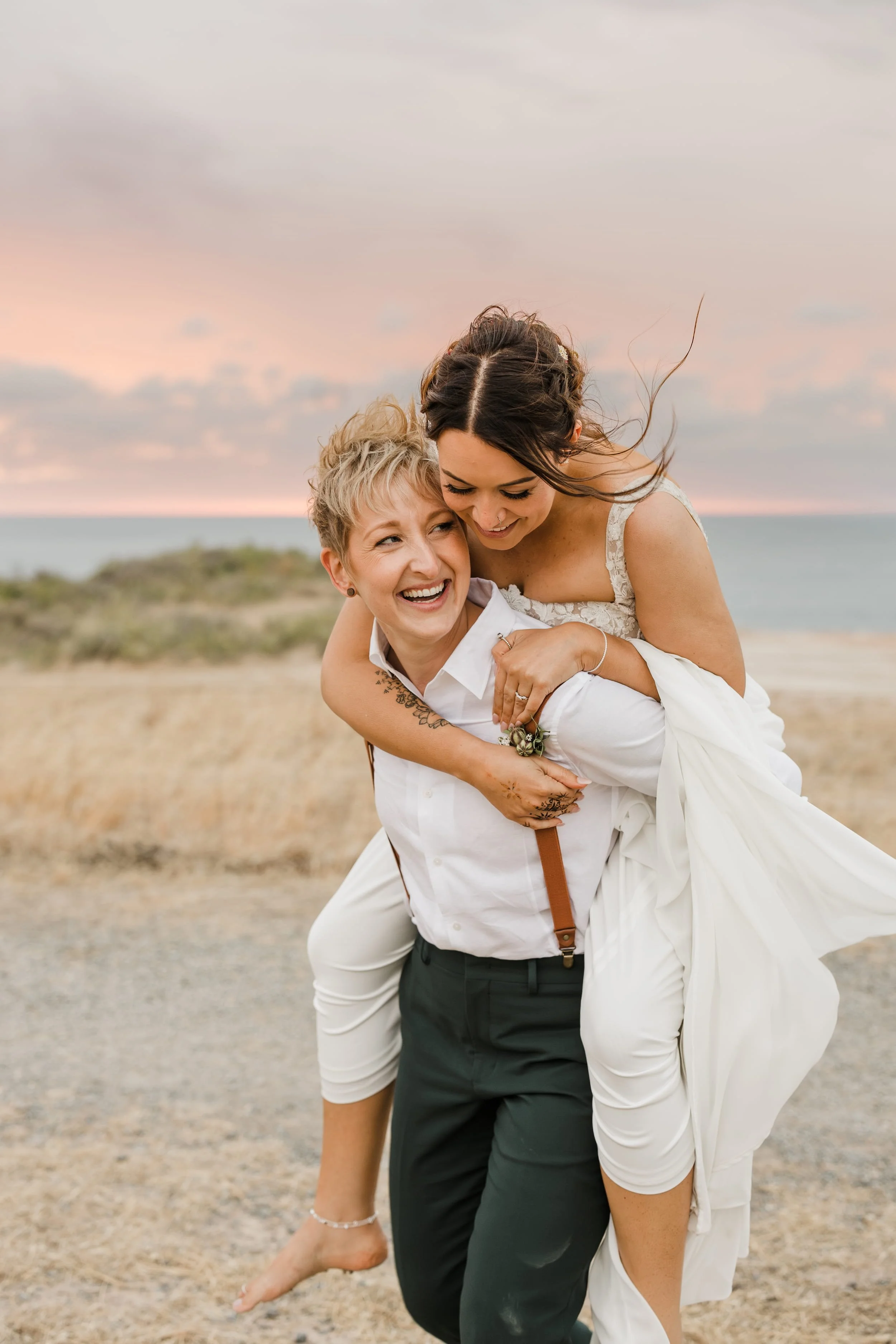 An Adelaide wedding photographer's photo of two bride's sharing a joyful moment outdoors with a beach and cloudy sky in the background. One woman is giving a piggyback ride to the other.