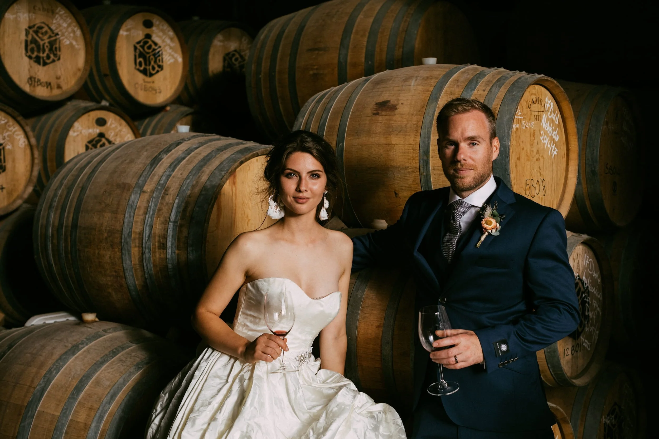 Bride and groom standing in a wine cellar among stacks of wine barrels, each with handwritten markings. The bride is holding a glass of red wine, and the groom is holding a glass of white wine. Both are dressed in wedding attire. The bride has dark hair styled up with earrings, and the groom is wearing a navy blue suit with a boutonniere.