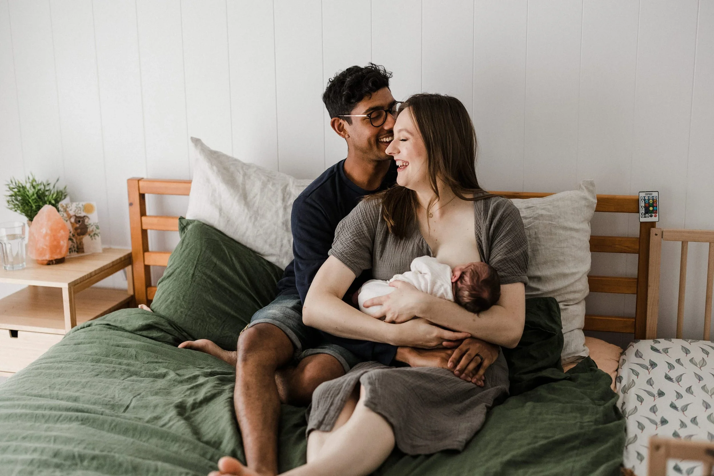 A professional portrait photo of a couple and their newborn baby, having a natural moment together laughing at the baby breastfeeds. The bed has a green cover, and the outfits match the colour palette well.