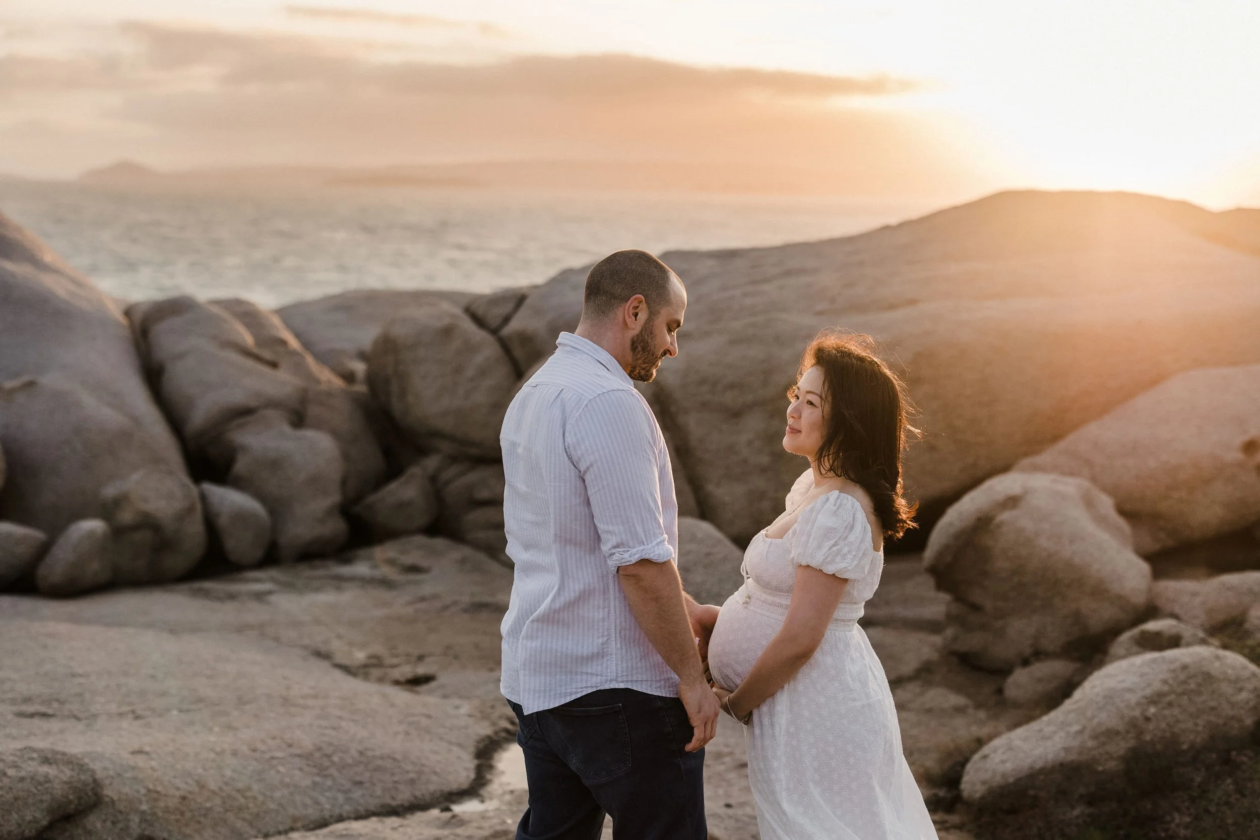 A couple holding hands and looking at each other on a beach at sunset with rocks and the ocean in the background.