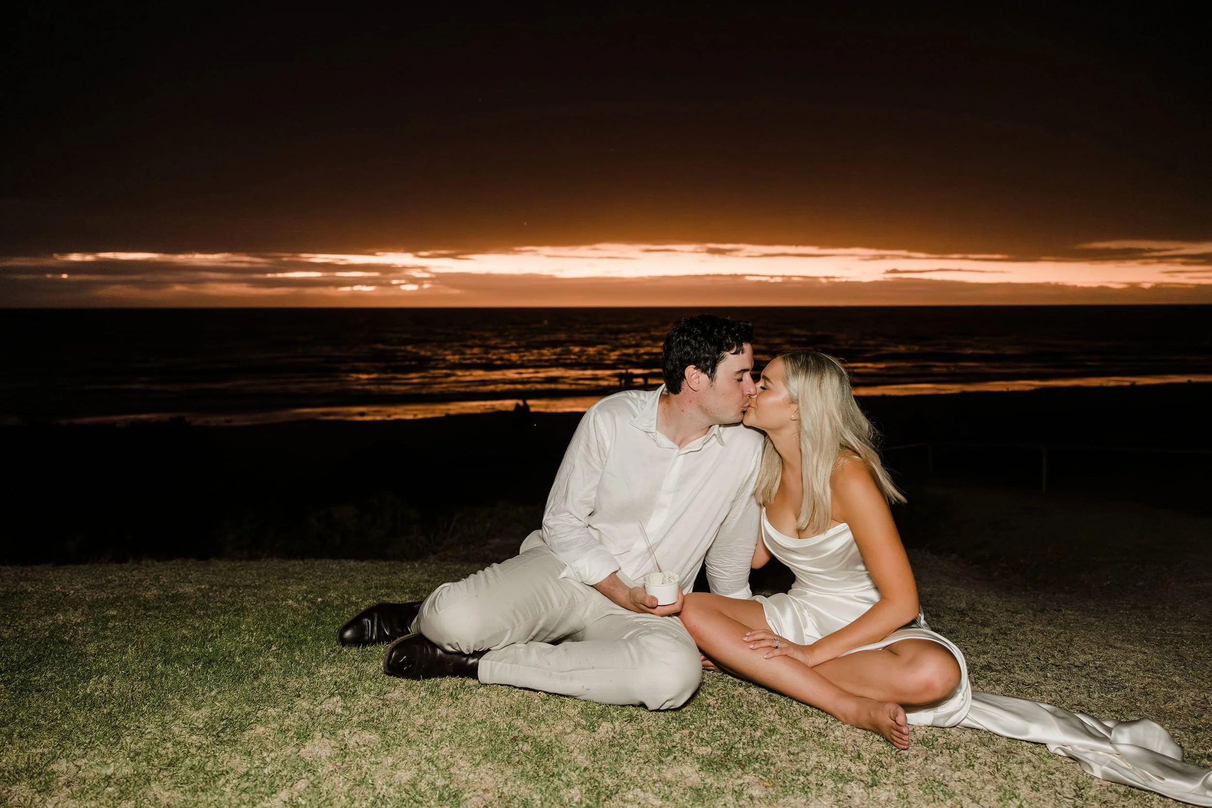 A couple dressed in white sitting and kissing on a grassy area near the beach during sunset or dusk, with a dark sky and orange clouds over the ocean.