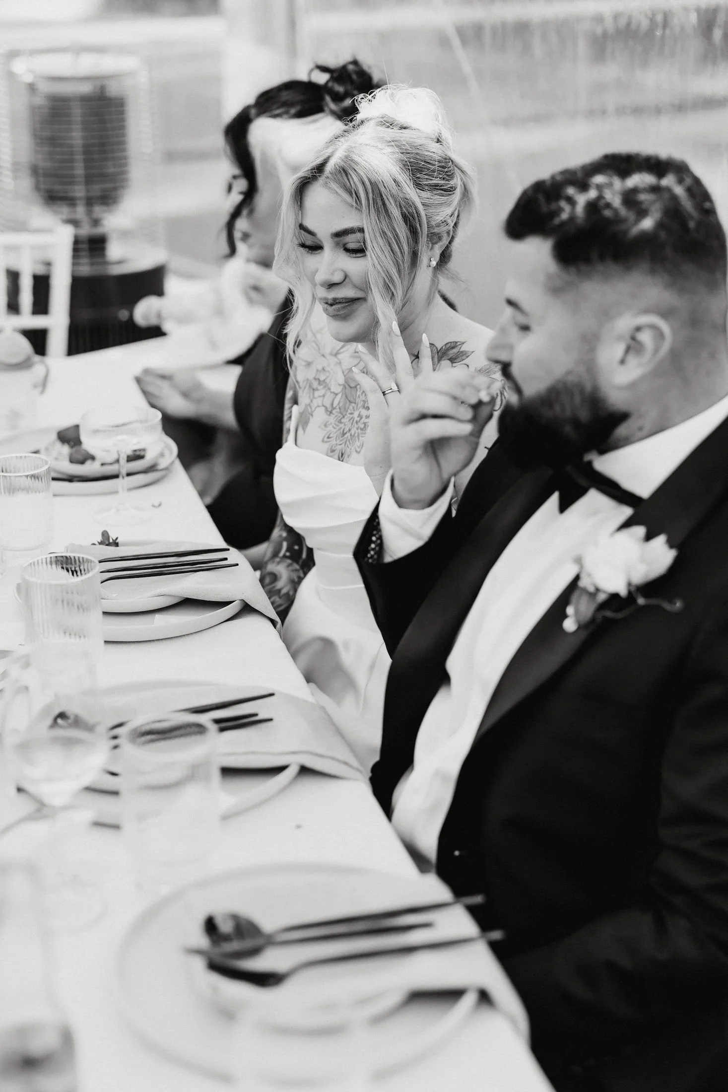 Black and white photo of a group of people sitting at a formal event, dressed in tuxedos and elegant dresses, enjoying a meal. The focus is on a woman with blonde hair, wearing earrings, and a man with dark hair and a beard, wearing a tuxedo with a bow tie. They are sitting at a table set with plates, glasses, and cutlery.