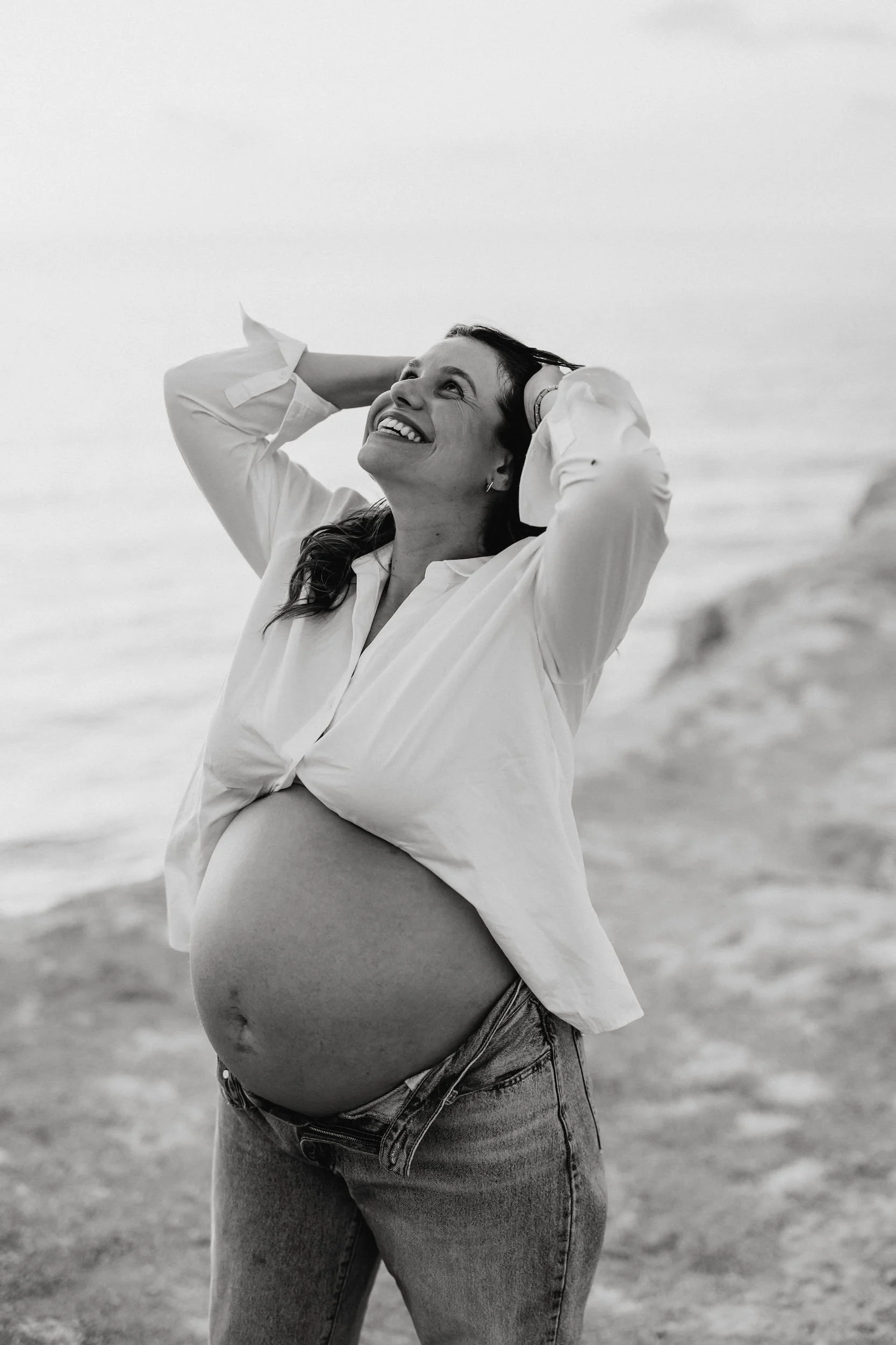 A pregnant woman with long dark hair, smiling, standing outdoors on a beach with a cloudy sky, wearing a white shirt and jeans.
