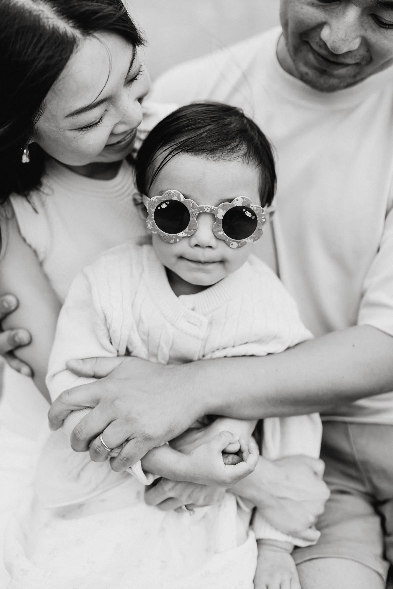 A black and white close-up photo of a young boy wearing glasses with a floral pattern and round lenses, being embraced by three adults, likely his parents, in a tender moment of affection.