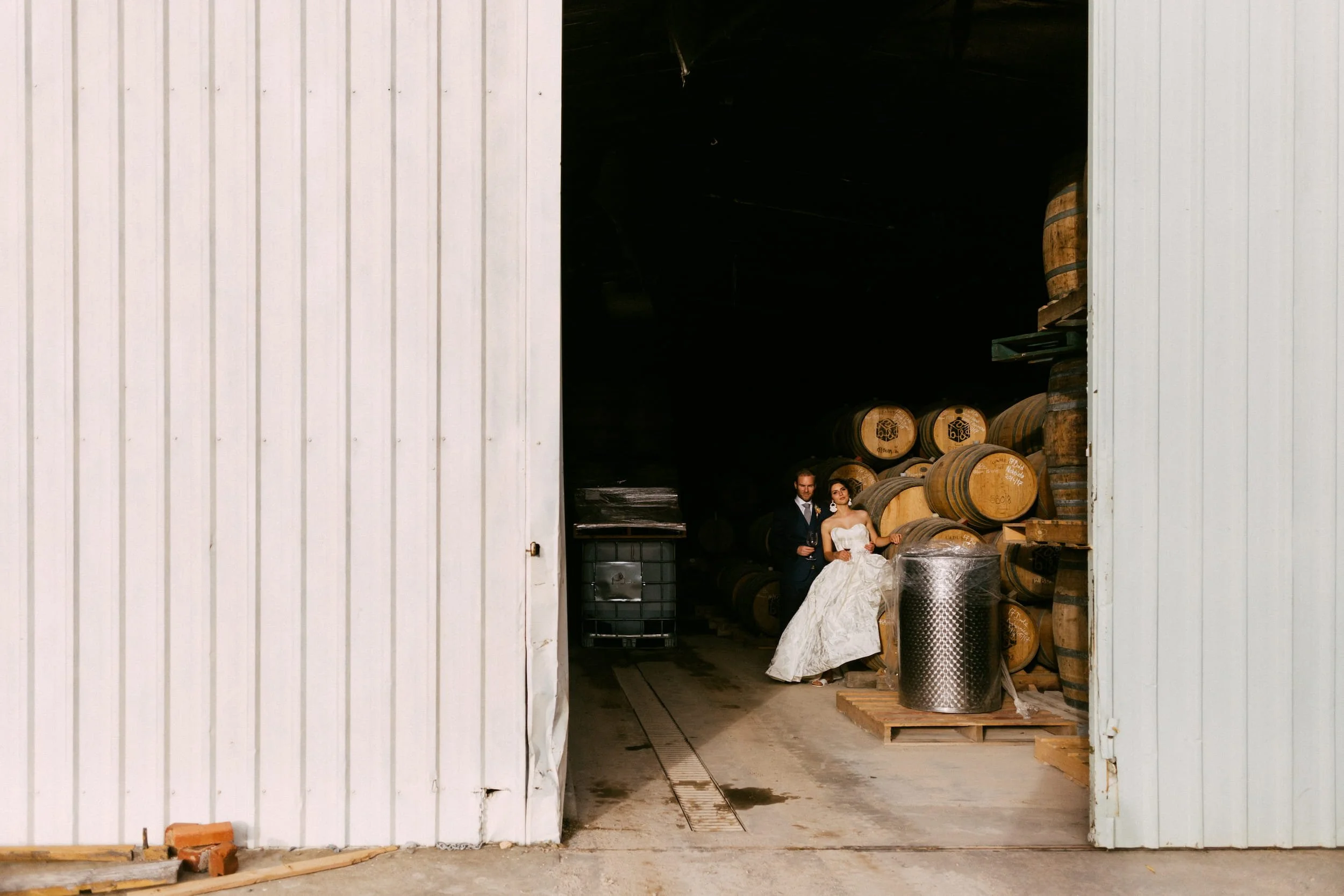 A bride and groom standing inside a warehouse with stacked wine barrels and industrial equipment, visible through an open large sliding door.