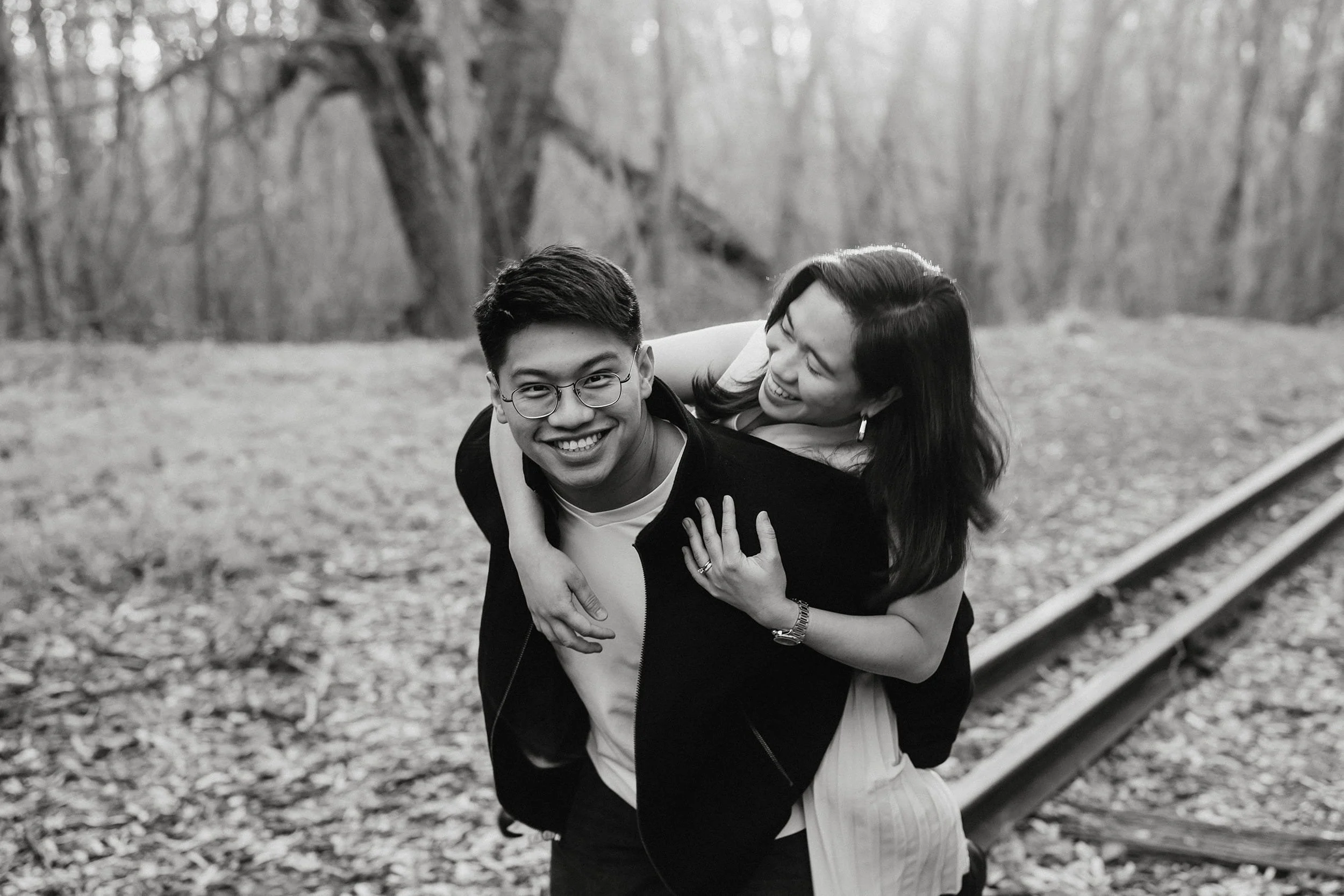 A black and white photo of a smiling young man giving a piggyback ride to a young woman, both outdoors in a wooded area.