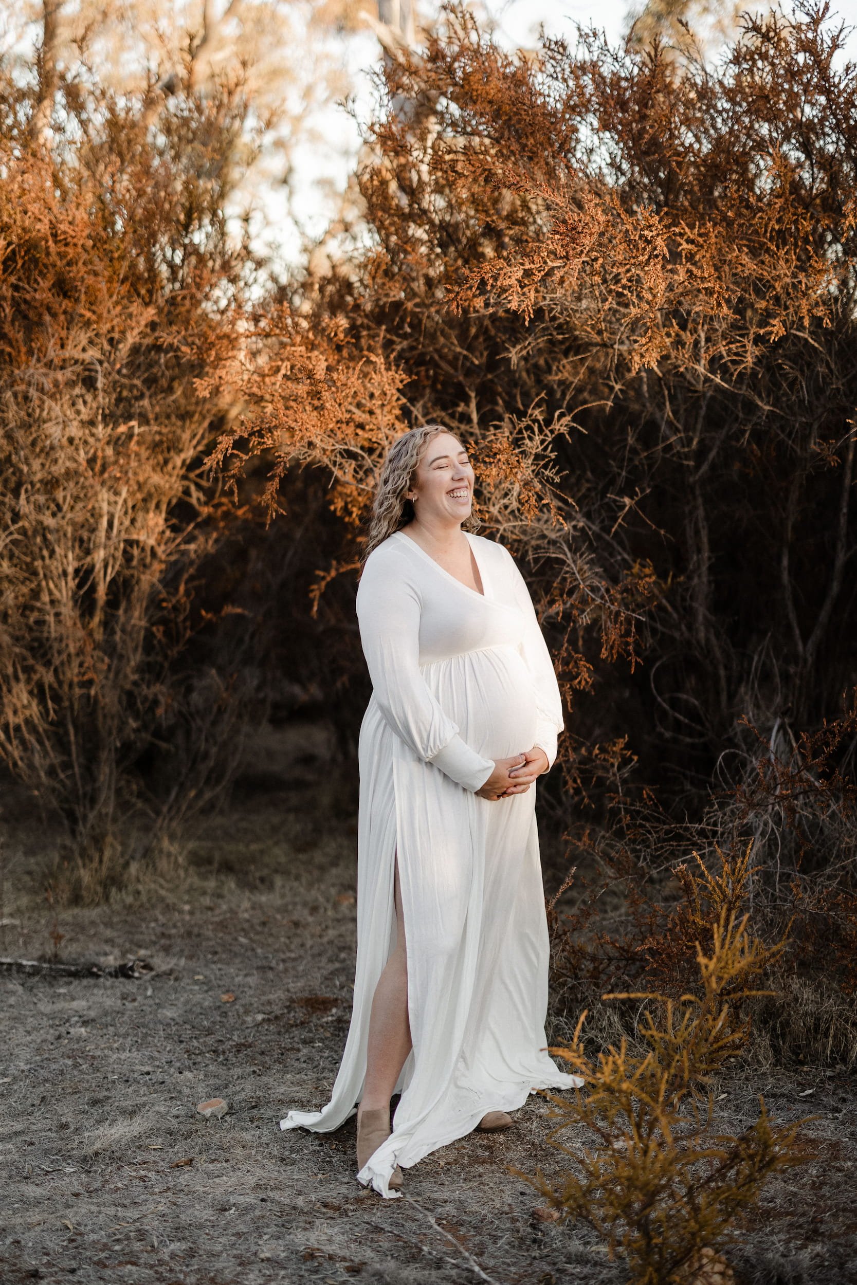 A pregnant woman in a white dress with a slit, standing outdoors among dry bushes with orange and brown foliage, smiling with her eyes closed and hands on her belly during sunset or golden hour.