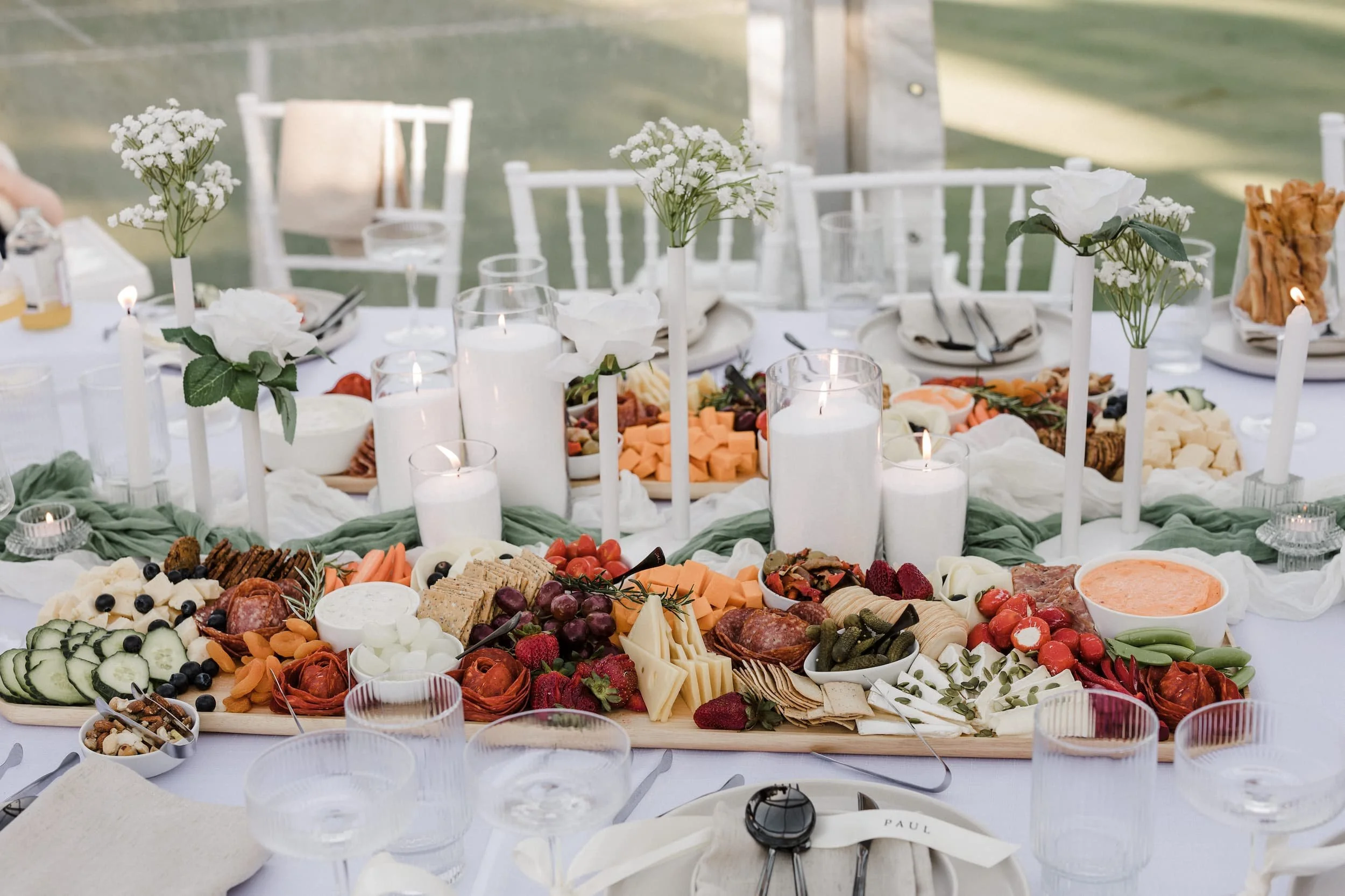 A table set with a cheese and charcuterie platter, fresh fruits, vegetables, and snacks, decorated with white candles and flowers, at an outdoor event.