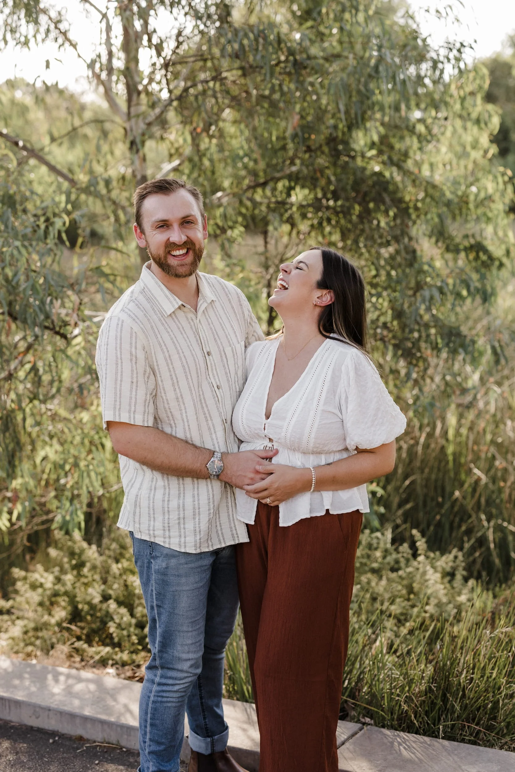 A happy couple standing outdoors near trees, smiling and laughing together, with the man wearing a striped shirt and jeans, and the woman wearing a white blouse and brown pants.