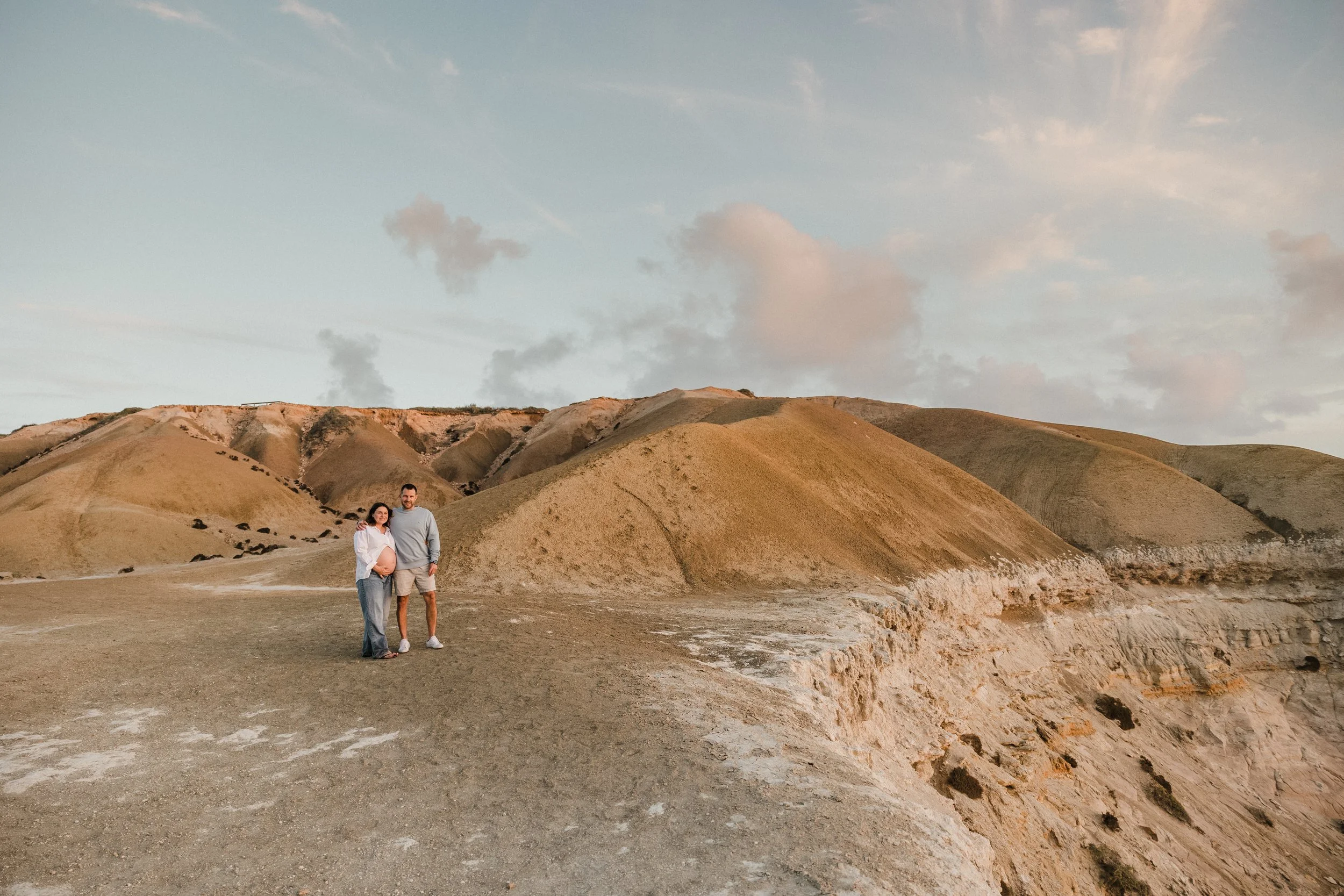 A professional maternity portrait photograph of a couple within a wide landscape. The colours within the outfit are reflected in the sky, and they stand out against the sandy hills.