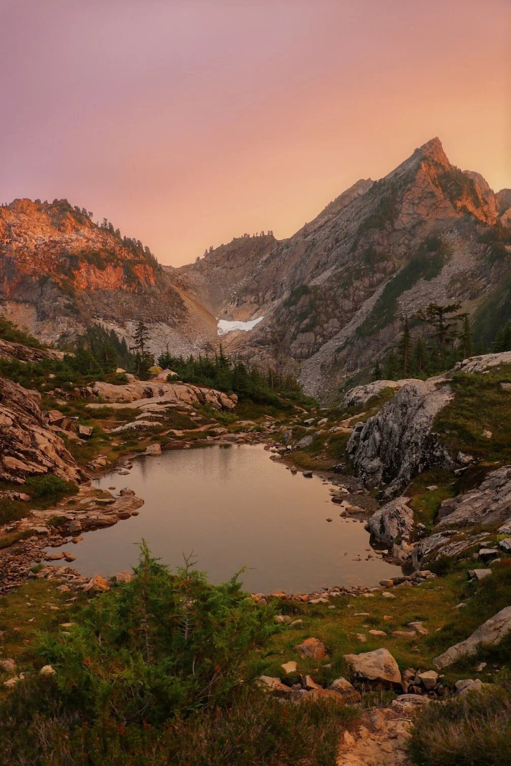 A mountainous landscape during sunset with rocky peaks, a small reflective lake, green trees, and a pinkish sky.