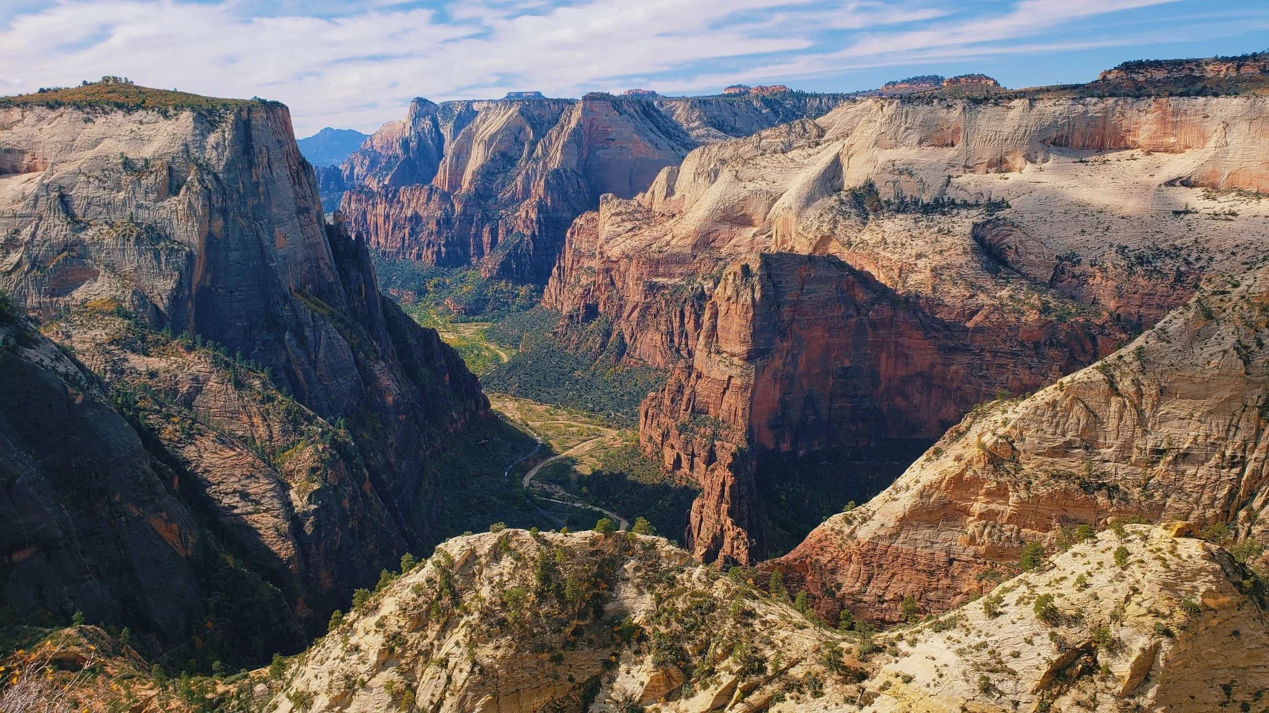 A panoramic view of a deep canyon with steep, layered rock walls and a winding river at the bottom, under a partly cloudy sky.