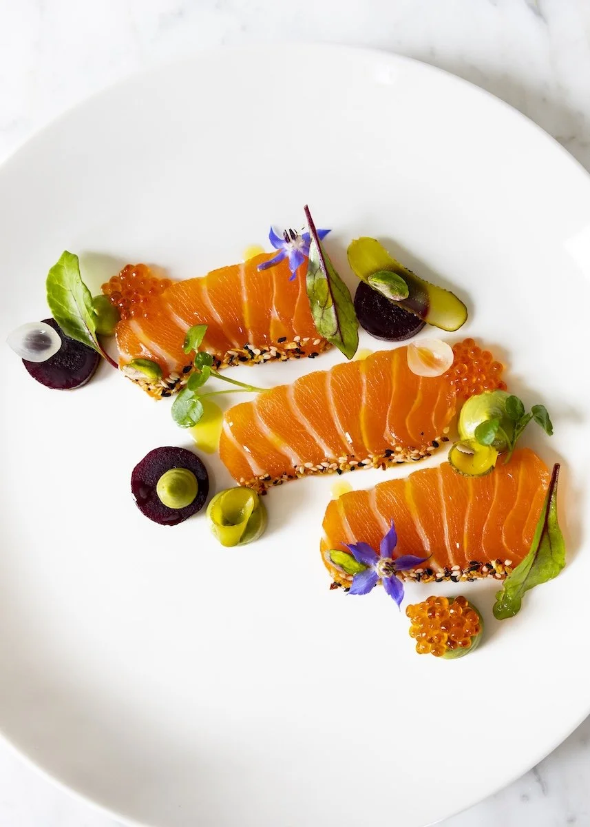 Sliced salmon sashimi garnished with microgreens, edible flowers, black sesame seeds, and various colorful garnishes on a white plate.