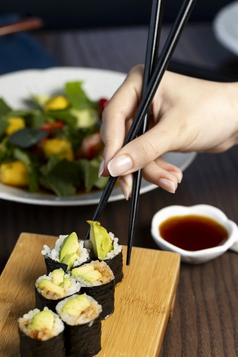Someone using chopsticks to pick up sushi rolls with avocado on top, on a wooden serving board, with a small dish of soy sauce and a salad in the background.
