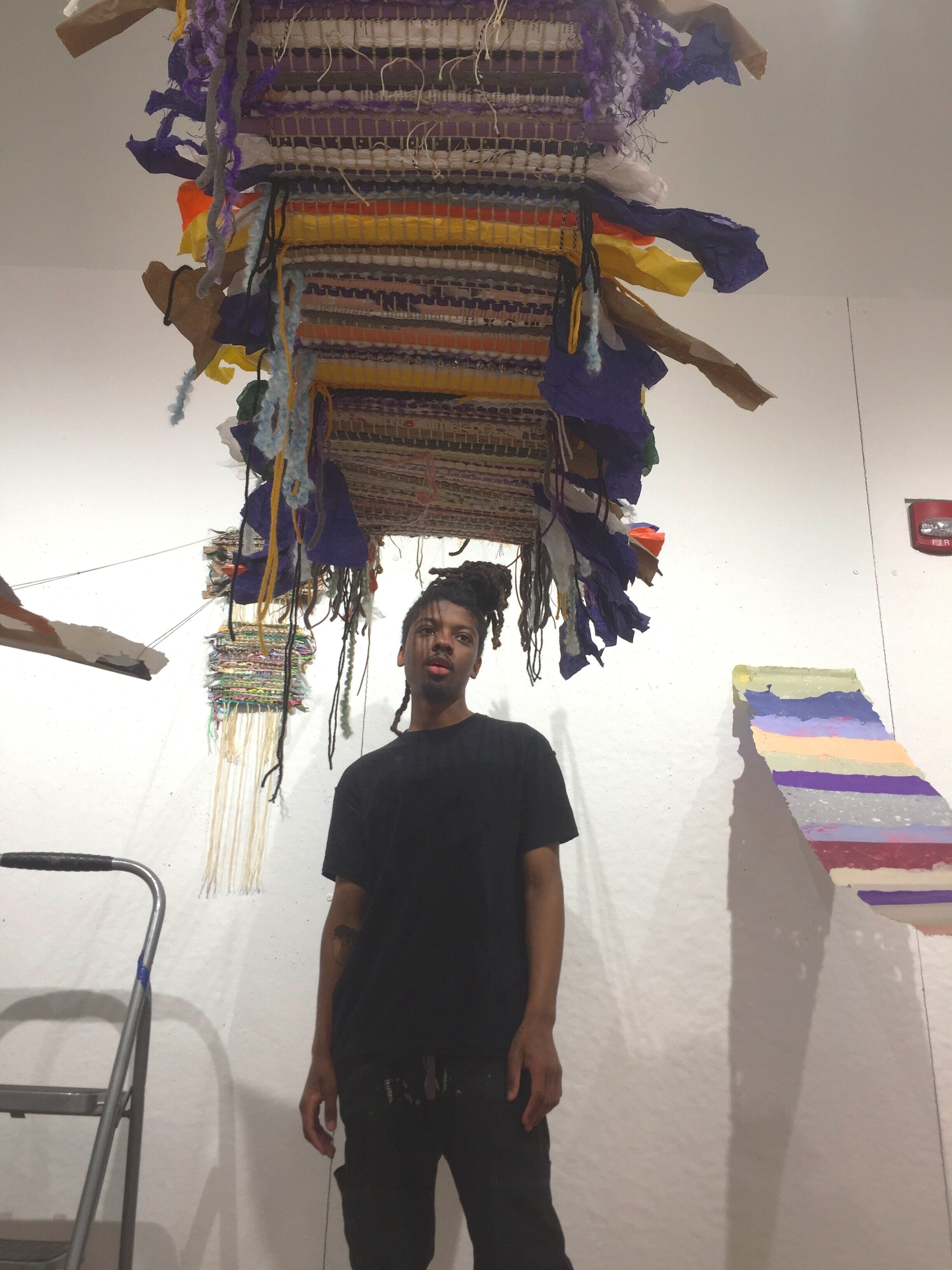   Student, Philly Johnson stands underneath the work of Andrea Romero, who created a mobile weaving studio for her train commute.  