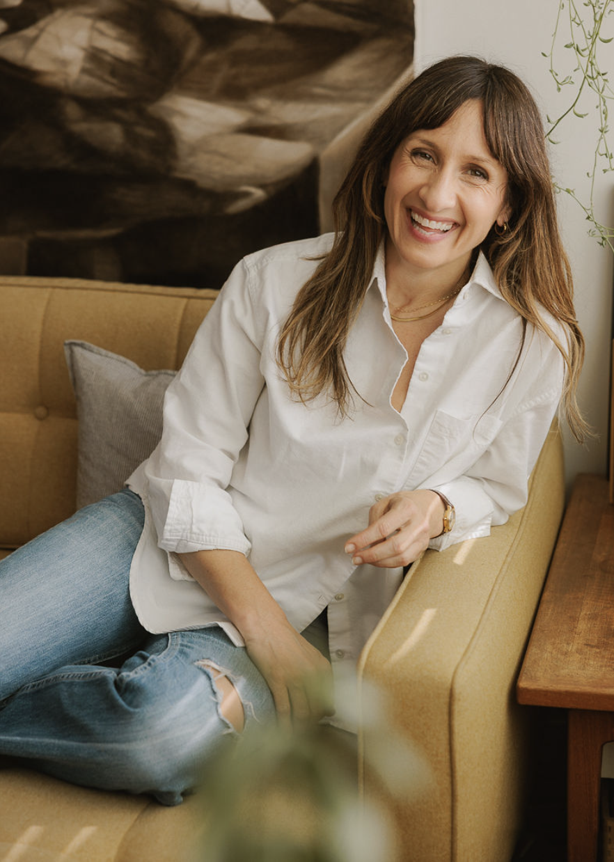 Amanda, with long brown hair is sitting on a beige couch, smiling and looking at the camera. She is wearing a white button-up shirt and ripped jeans. There is a gray pillow behind her and a wooden side table nearby.