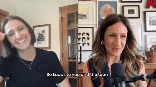 Two women sitting in separate spaces during a video call, one laughing and the other speaking into a microphone, with a background of framed art and photos.