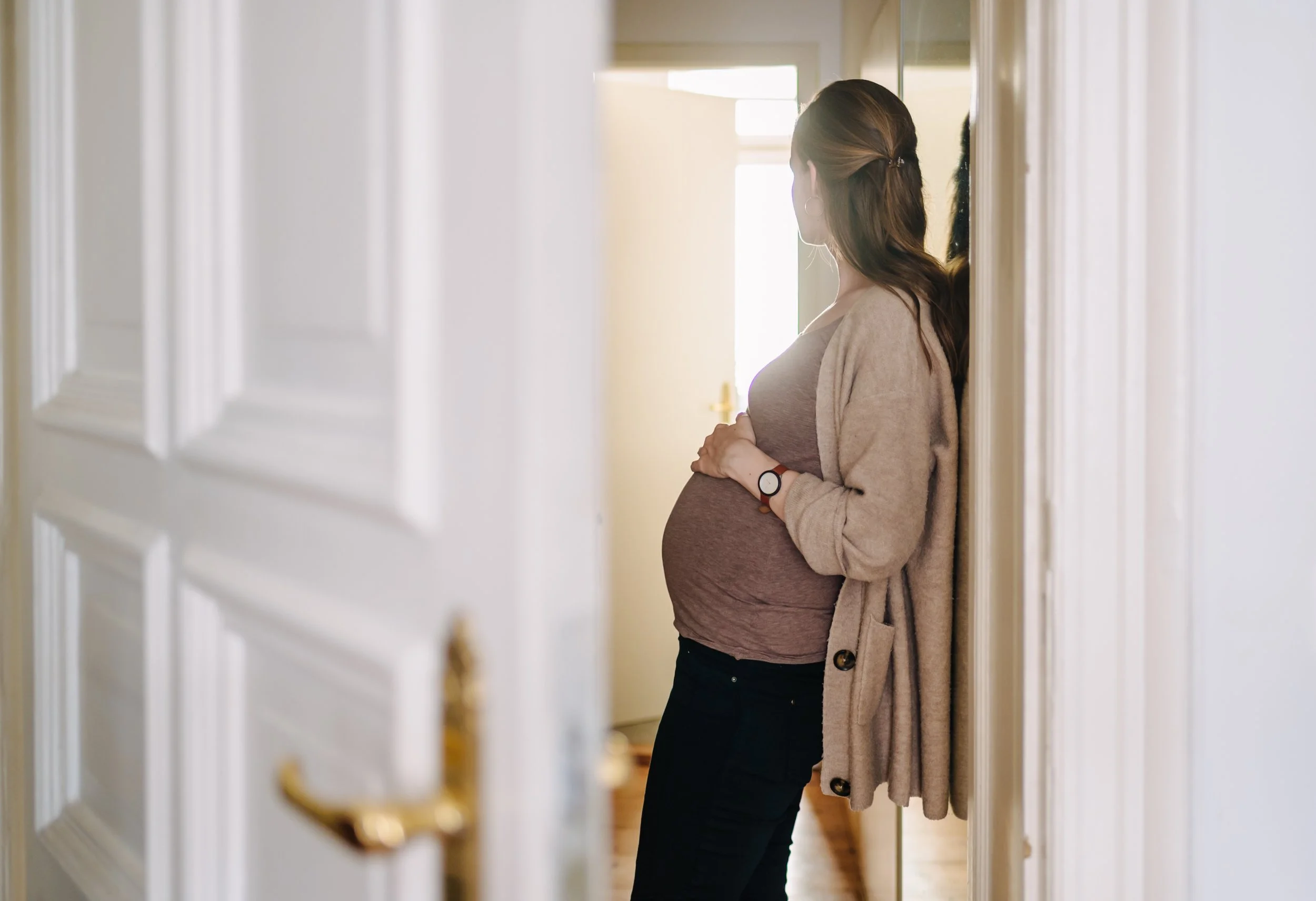 Schwangere Frau lehnt mit den Händen auf dem Babybauch im Flur und fotografiert sich selbst.