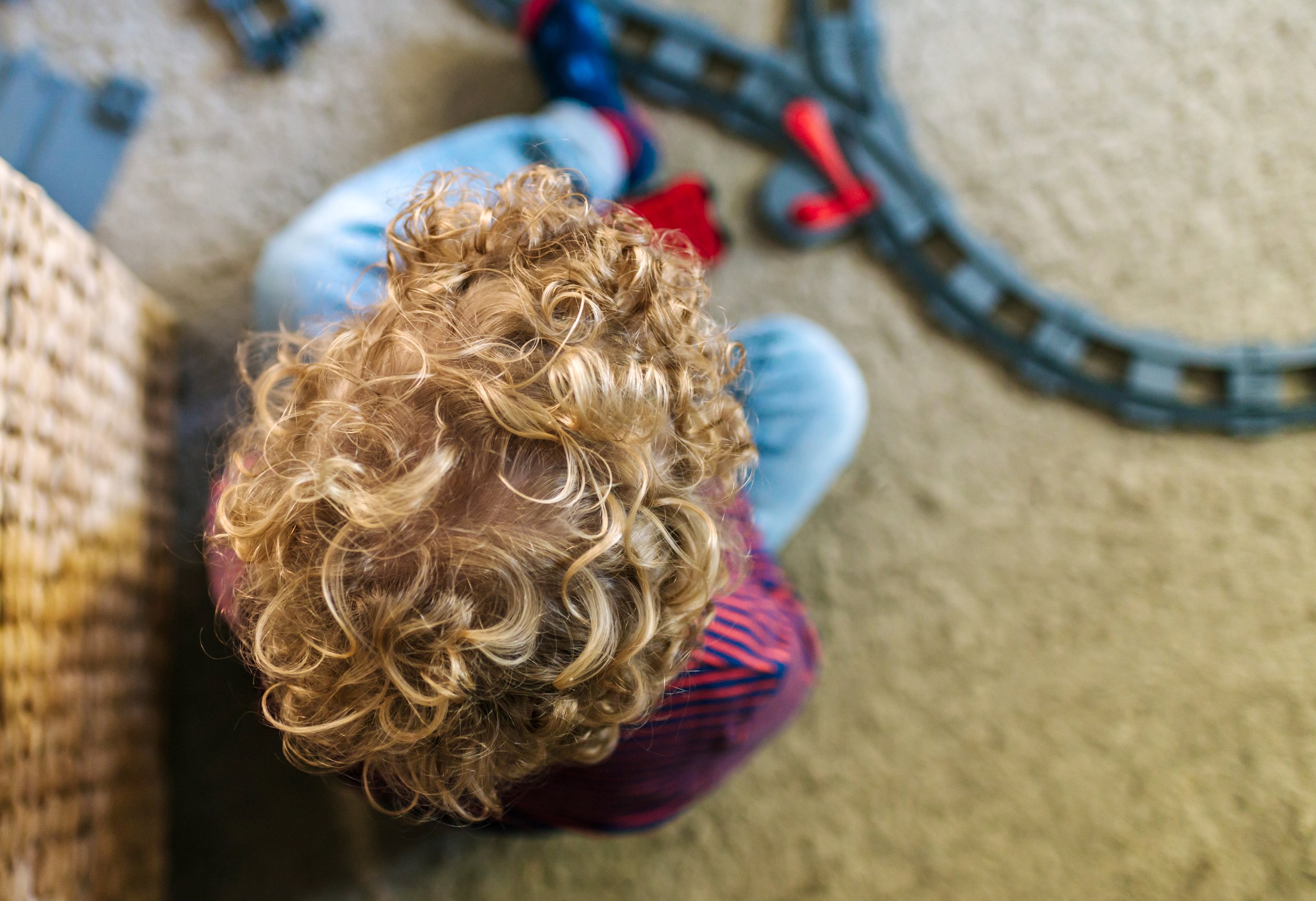 Man sieht ein Kind von oben mit blonden Locken, das auf dem Teppich mit einer Eisenbahn spielt.