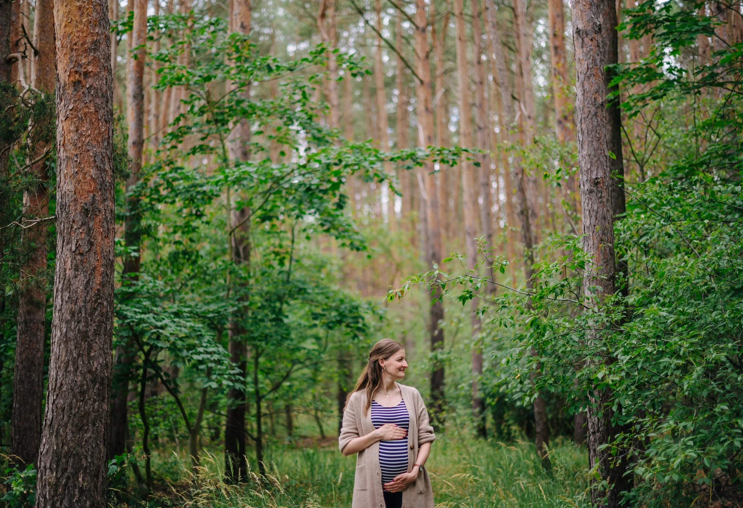 Schwangere Frau mit gestreiftem Oberteil fotografiert ihren Babybauch im Wald.