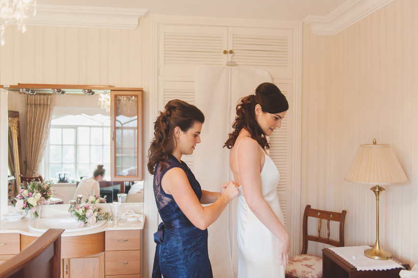 Bride's sister doing up her wedding dress on the morning of the wedding in a cream bedroom
