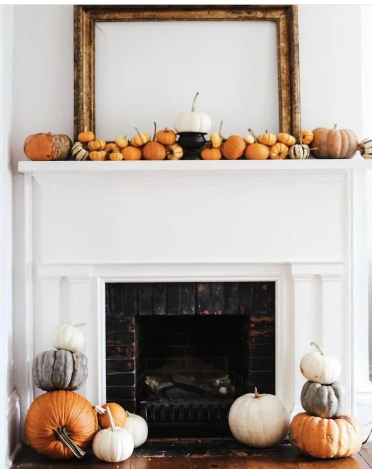 White fall fireplace mantel with an empty antique gold frame, a row of orange and white mini pumpkins, and stacked heirloom pumpkins on the hearth