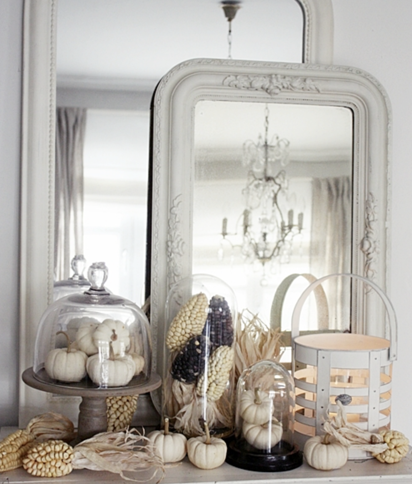 All-white French country fall mantel with layered ornate mirrors, glass cloches over mini white pumpkins, dried Indian corn, and a white lantern