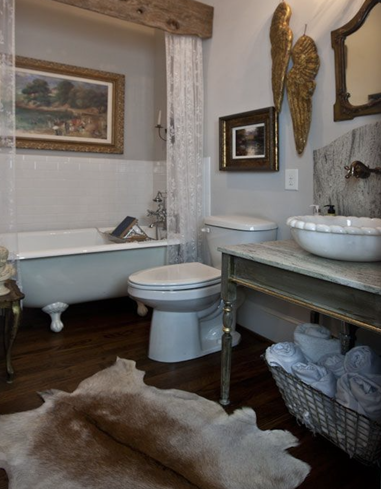 Old-world western bathroom with wood beam over clawfoot tub, gilded landscape art, antique vanity, and cowhide rug