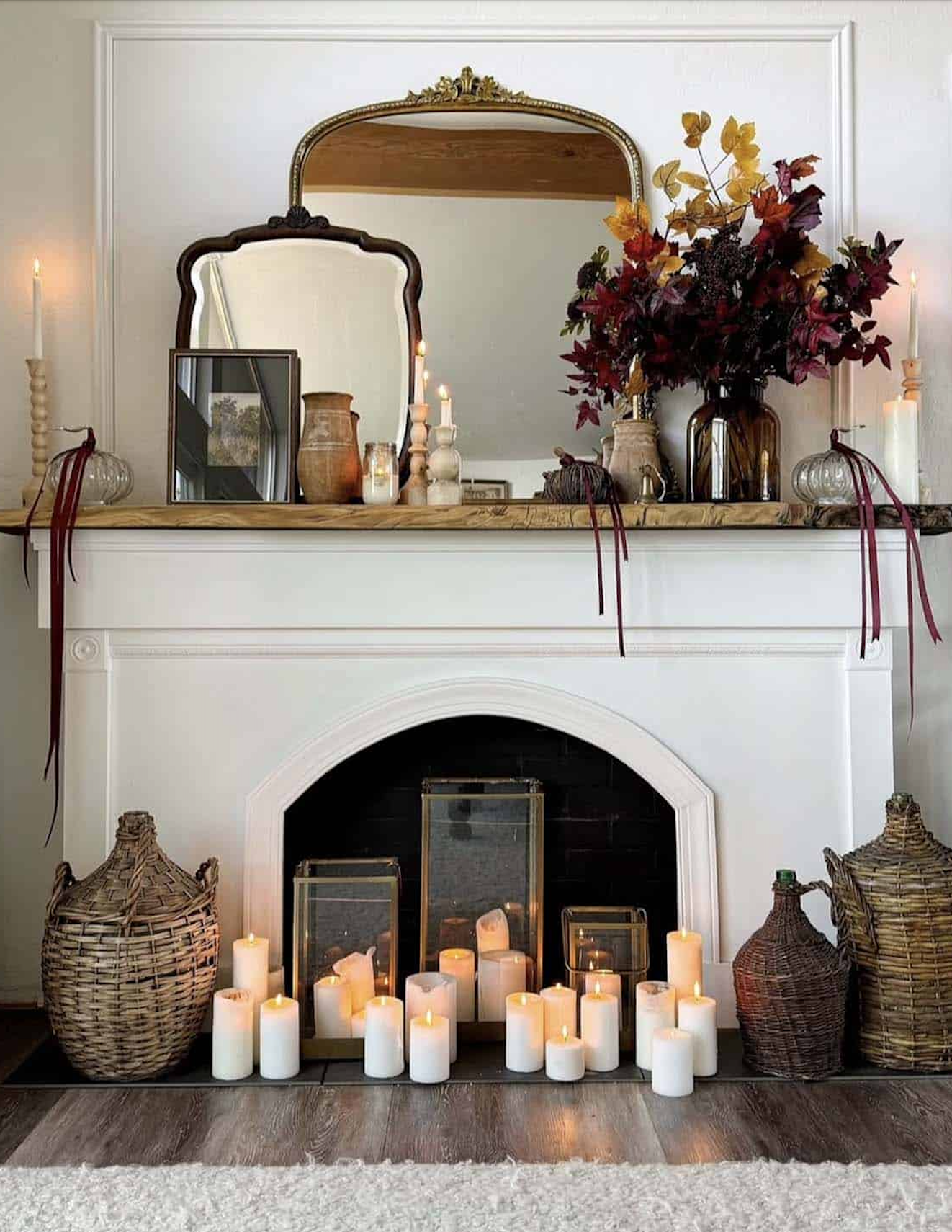 European cottage fall fireplace with a live-edge wood mantel, layered vintage mirrors, an autumn leaf bouquet in an amber vase, glass pumpkins with burgundy ribbons, and pillar candles filling the firebox