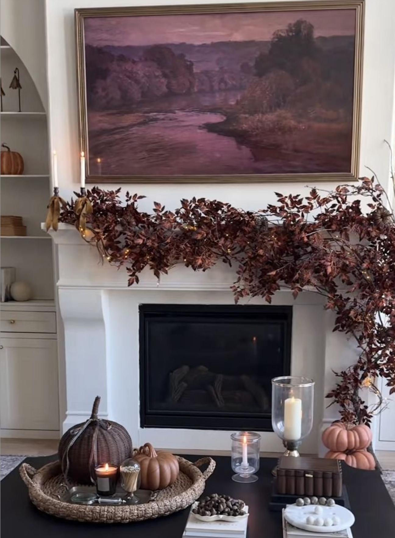 Elegant fall fireplace mantel with cascading burgundy leaf garland, fairy lights, and a plum landscape painting in a gold frame