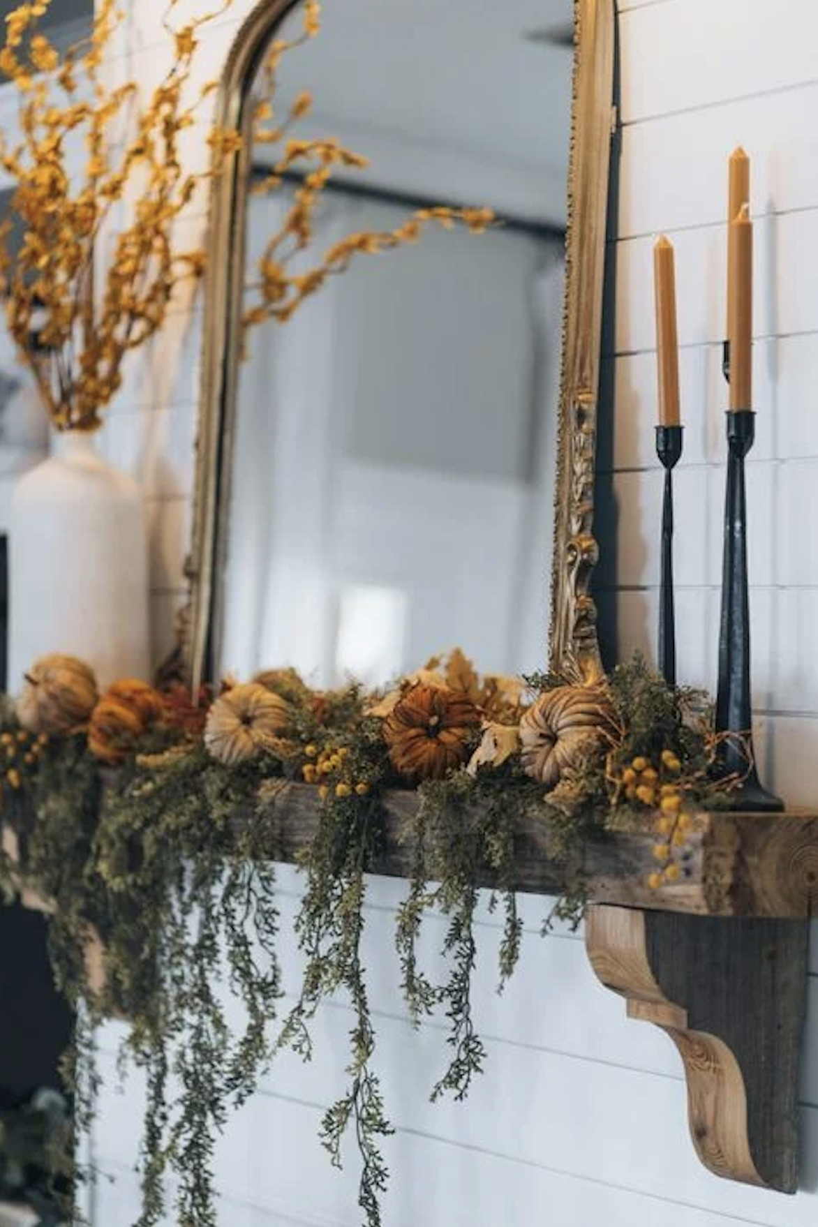 Rustic wood beam fall mantel with velvet pumpkins, trailing green garland, yellow billy button flowers, a gold arched mirror, and black candlesticks with mustard taper candles