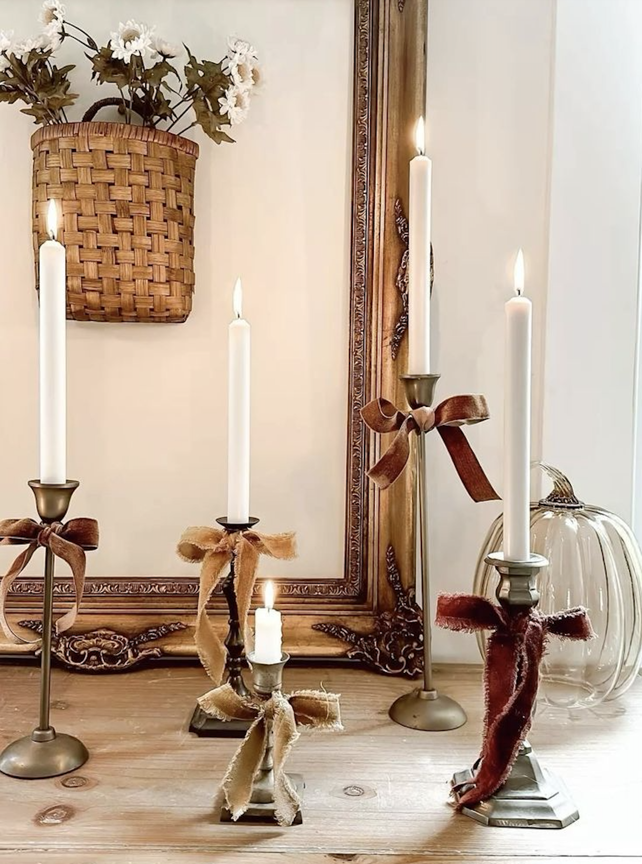 Mismatched brass candlesticks with white taper candles tied with rust, mustard, and burgundy ribbon bows, beside an empty gold frame, a clear glass pumpkin, and a woven basket of white flowers