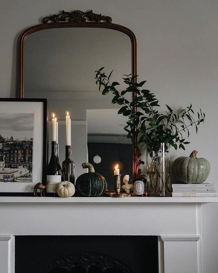 Moody fall mantel with an ornate bronze mirror, wine bottle candle holders, green and white pumpkins, a black and white city print, and greenery in a glass vase