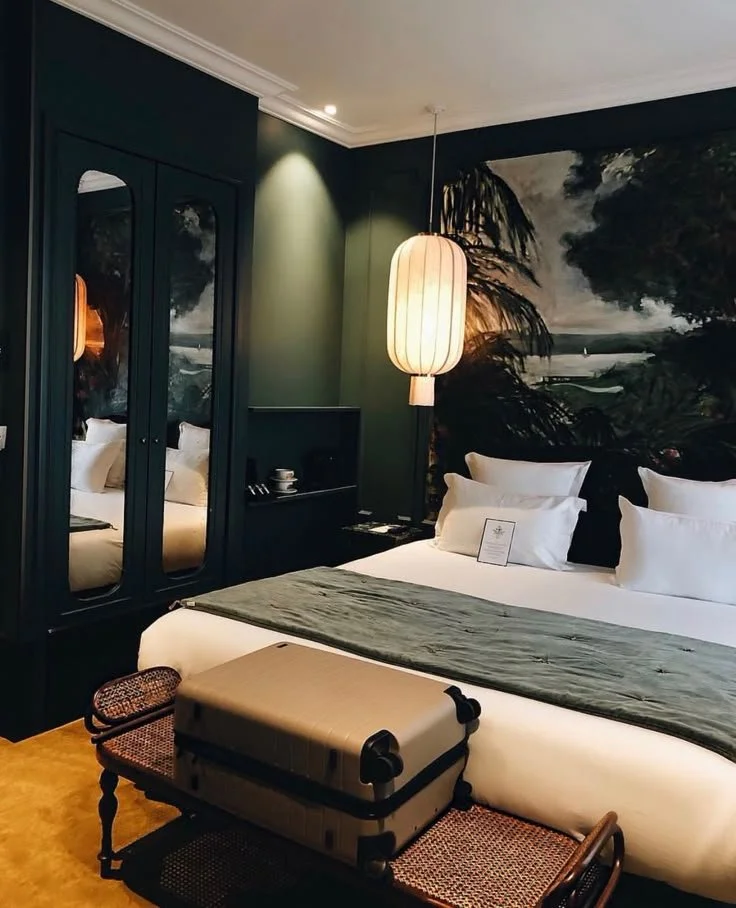 Dark green boutique hotel bedroom with a painted landscape mural behind the bed, a paper lantern pendant, mirrored arched wardrobe doors, a sage velvet throw and a cane luggage bench.