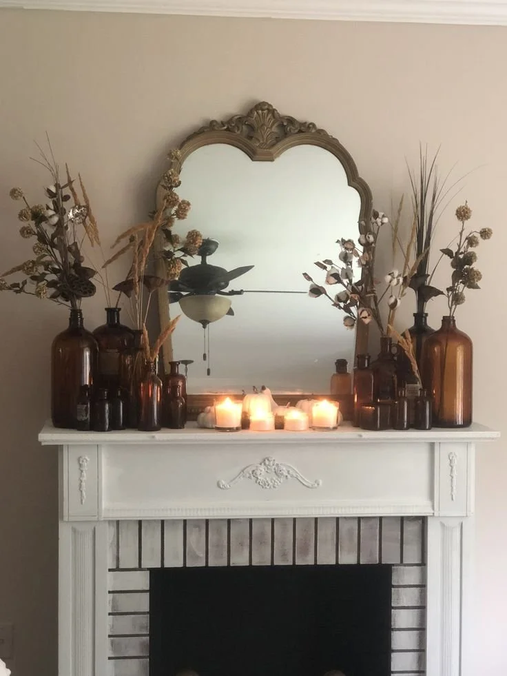 White fall fireplace mantel with a collection of amber glass apothecary bottles, dried cotton stems, wheat, and lotus pods, with glowing votive candles and a gold arched mirror