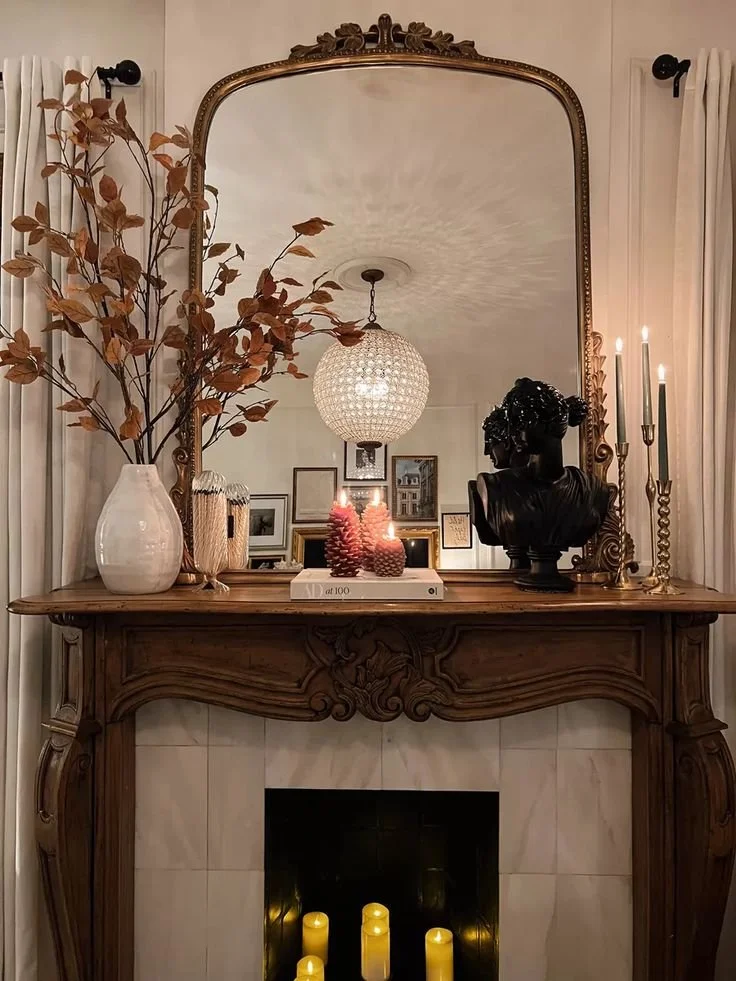 Carved French wood fireplace mantel with an ornate gold mirror, rust fall leaf branches in a white vase, pinecone candles, a black bust sculpture, and candles glowing in the firebox