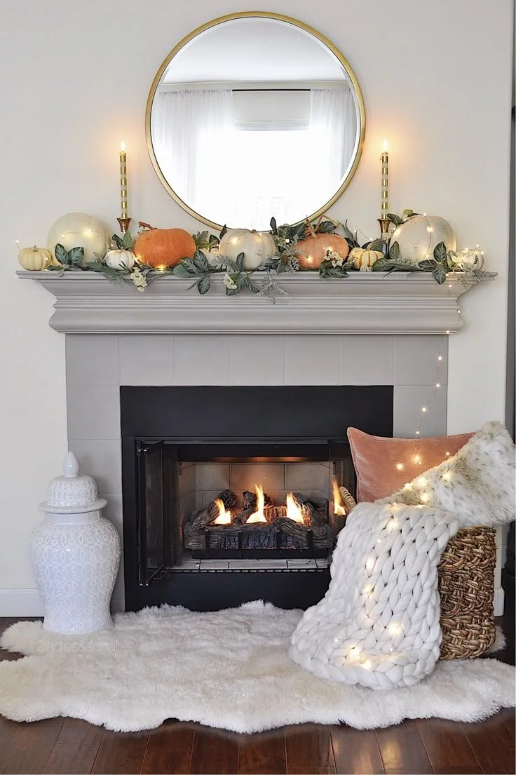 Cozy fall fireplace with a round gold mirror, white and orange pumpkins, greenery garland with fairy lights, and a chunky knit blanket in a basket on a faux fur rug
