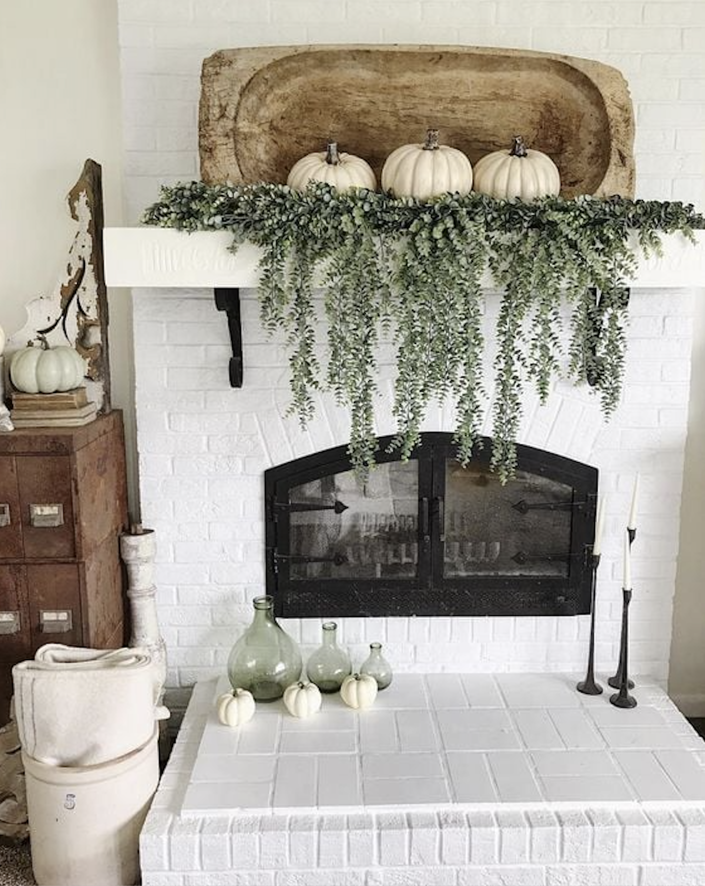 White brick fall fireplace with an antique dough bowl holding white pumpkins and a trailing eucalyptus garland cascading over the mantel