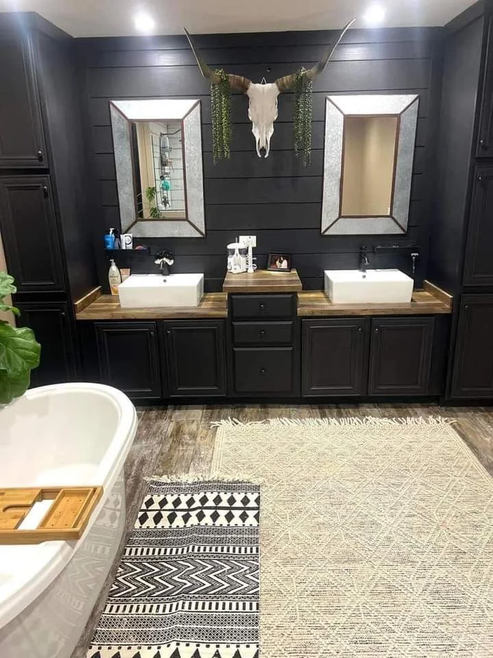 Black western primary bathroom with layered rugs, double vanity, galvanized mirrors, and longhorn skull with greenery