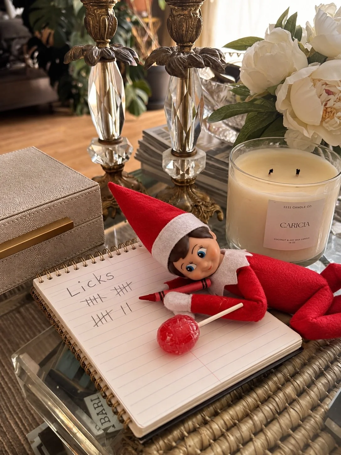 Elf on the Shelf asleep on a notepad with lollipop lick tally marks, a red crayon and a red lollipop