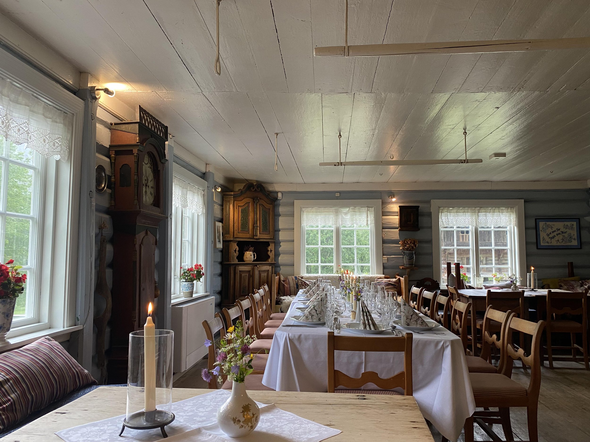 Innendørs rom med langbord dekket for selskap, store vinduer med gardiner, gamle møbler, blomster og lysestaker.