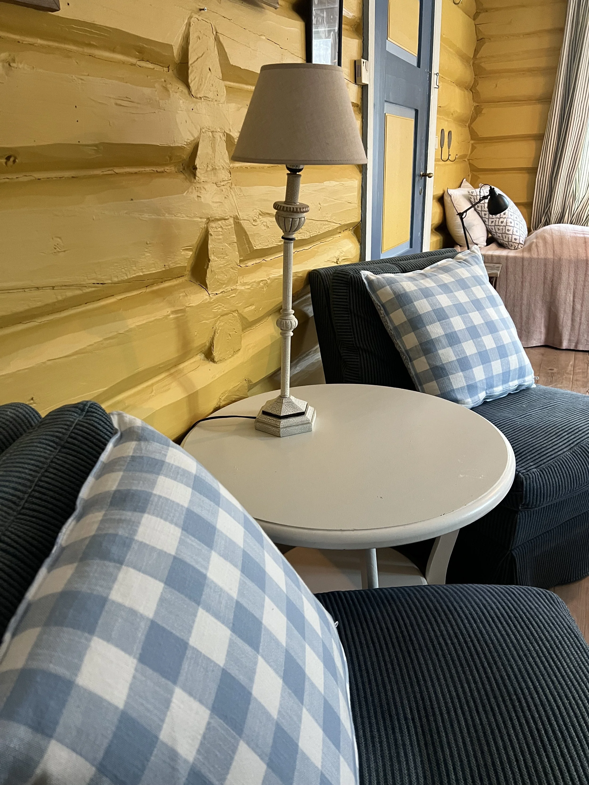 Interiør av et rom med stueinnredning, inkluderer et rundt hvitt bord med blå og hvite kryssende puter på en lenestol og sofa, en gul vegg med blå dører, en bordlampe, og vinduer med gardiner.